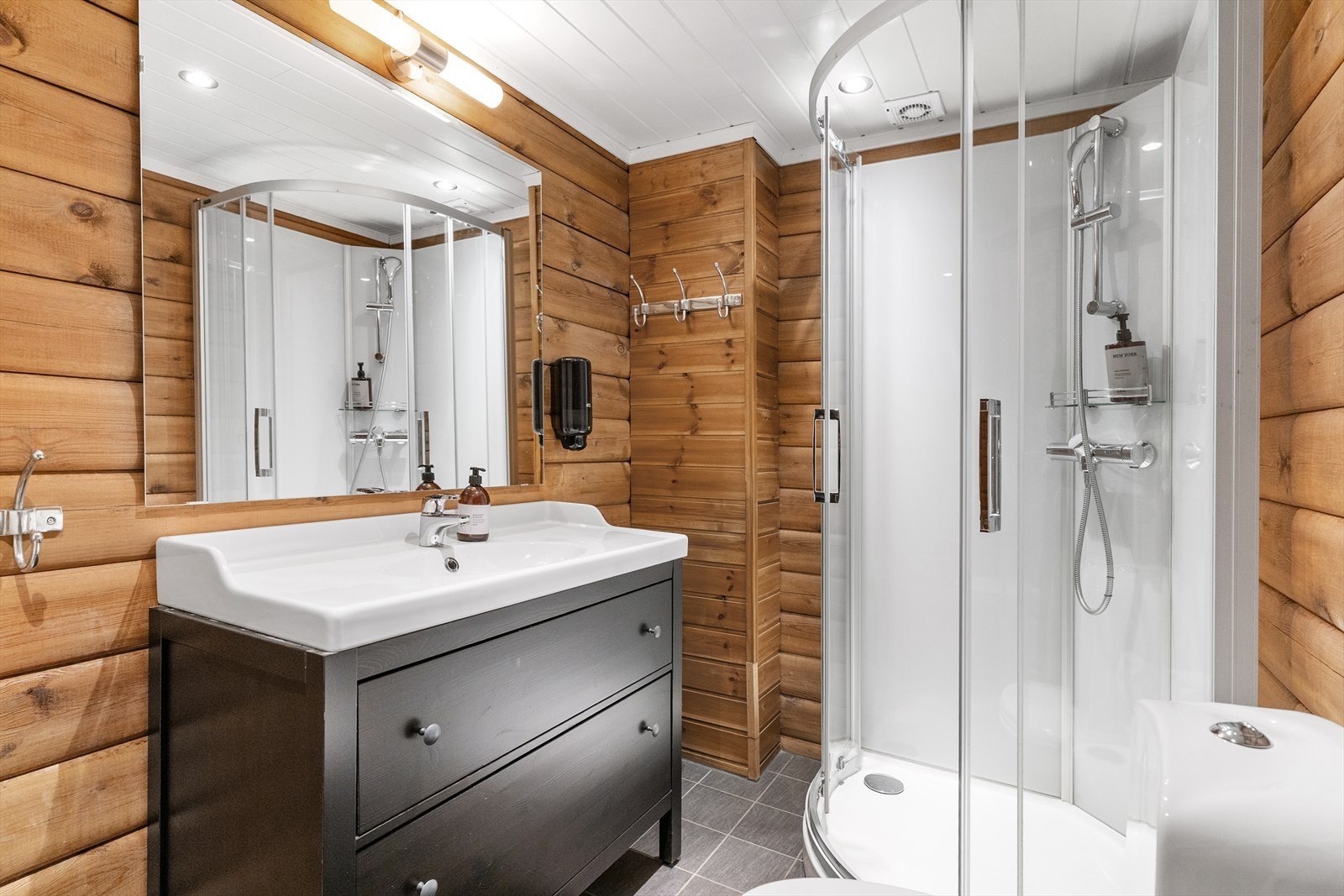 Badet inneholder dusjkabinett, servant med innredning, opplegg for vakemaskin og toalett. Vegger er av tømmer/laft, malt himling og gulv er fliselagt med elektriske varmekabler. Galleribilde