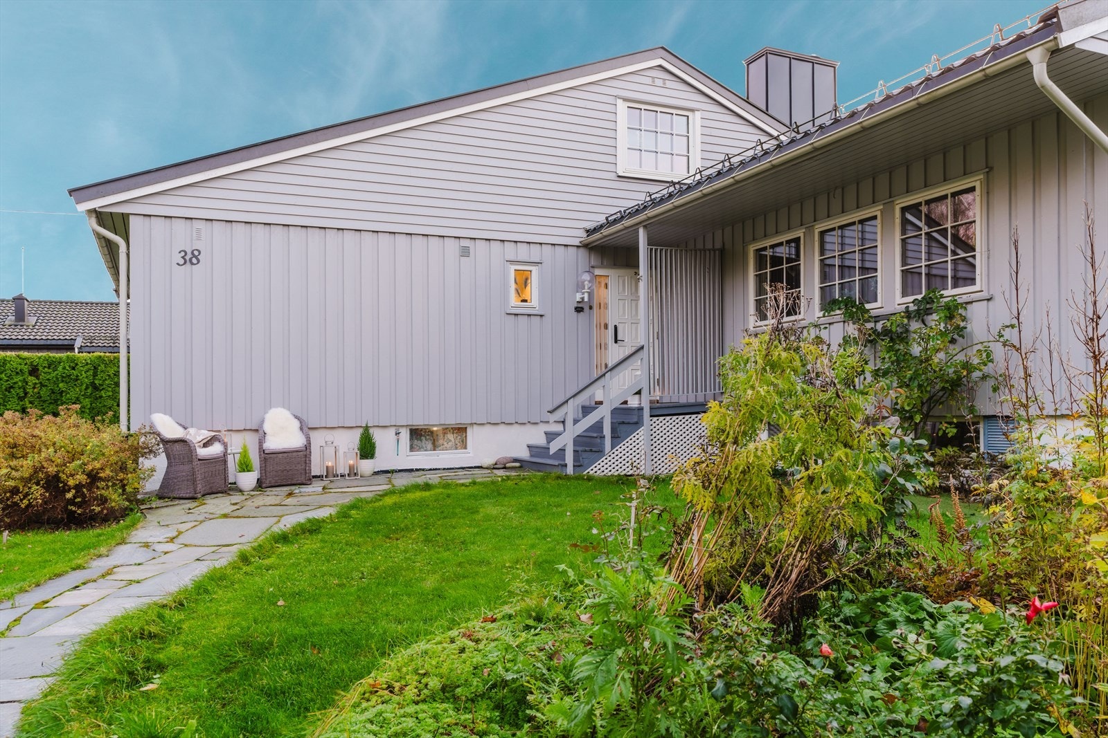 Inngangspartiet har en trapp i tre, og hagen er pent opparbeidet med blomsterbed. Galleribilde