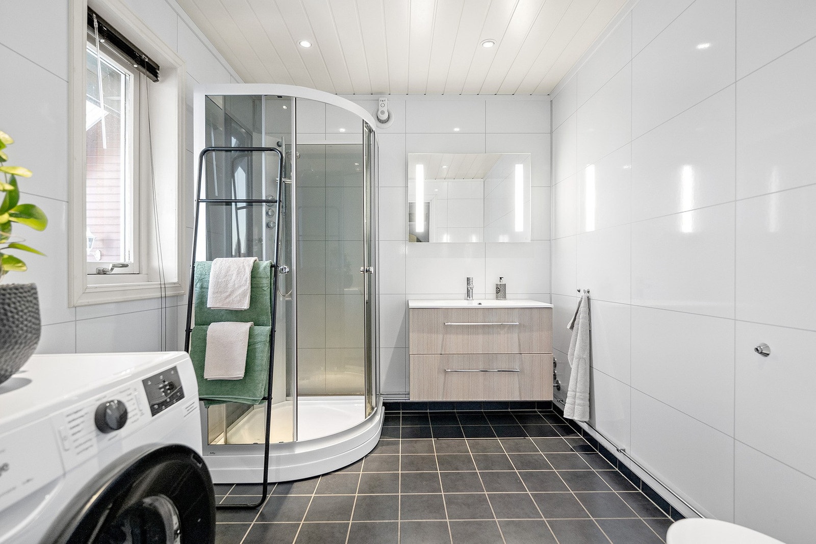 Badet i første etasje fremstår lyst, ryddig og funksjonelt med innredning bestående av servantskap, speil med lys, dusjkabinett, toalett og opplegg for vaskemaskin. Galleribilde