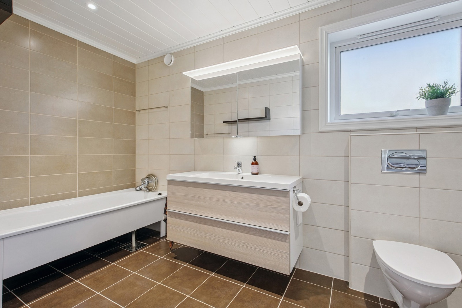 Rommet er innredet med nedfelt servant, veggmontert toalett, badekar og dusjvegger/hjørne Galleribilde