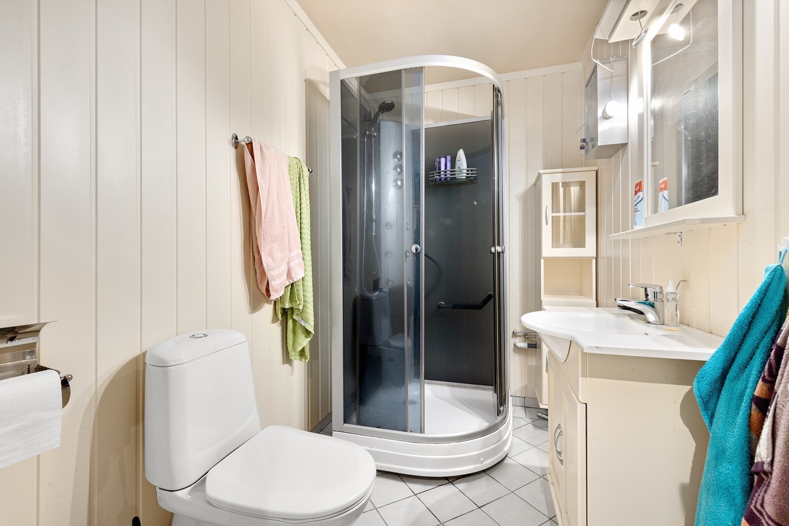 Baderom med varmekabler i flislagt gulv. Badet inneholder servantinnredning, vegghengt høyskap, frittstående toalett og dusjkabinett. Galleribilde