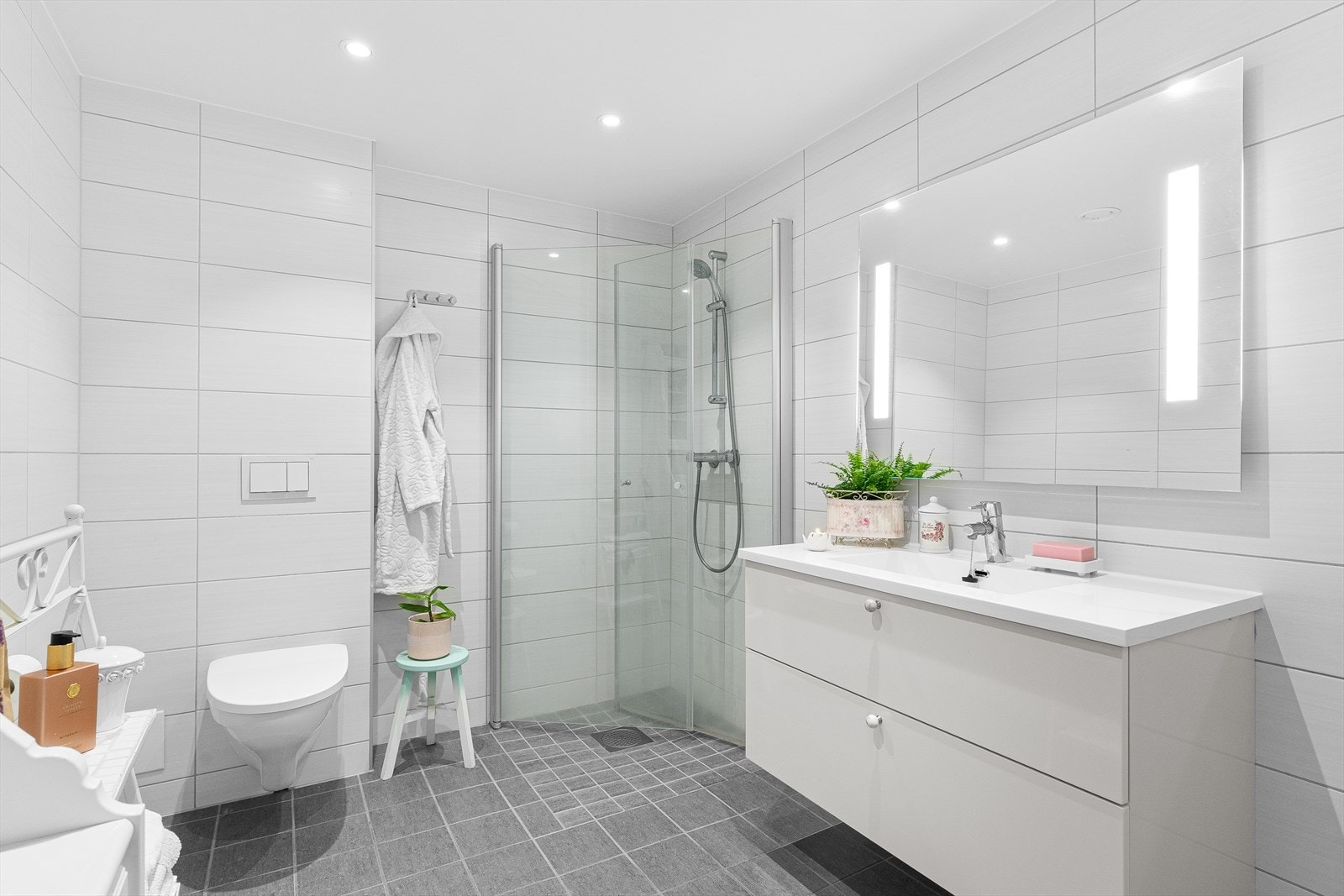 Lyst og pent flislagt bad med heldekkende servant i skuffinnredning, dusj på vegg med glassdører, og vegghengt toalett. Galleribilde