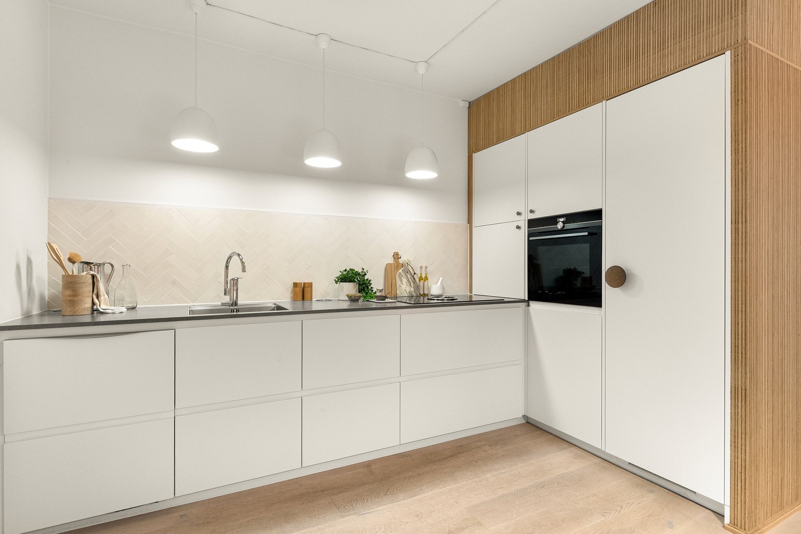 Kjøkkeninnredningen hvite glatte fronter, benkeplate med nedfelt kum i rustfritt stål, lekkert flisfelt i fiskebeinsmønster på vegg over kjøkkenbenk. Galleribilde