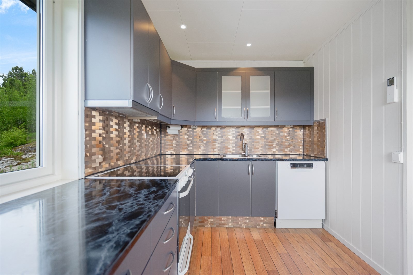 Ventilator i overskap og dekorplater med flisemønster på vegg over benk. Plass for komfyr, oppvaskmaskin og kjøleskap. Galleribilde