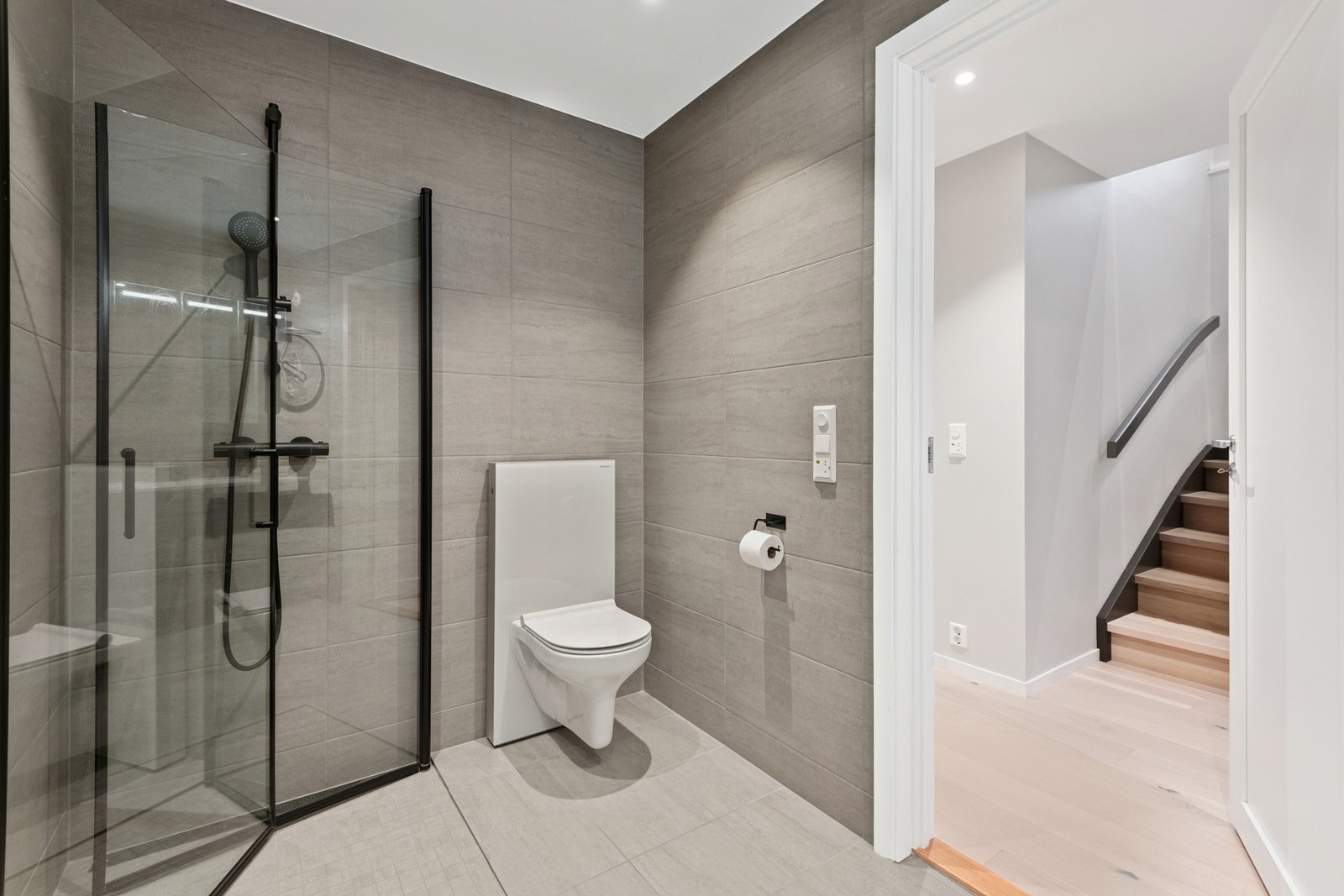 Badet har vegghengt toalett, flislagt gulv med varmekabler og dusjhjørne med sorte detaljer. Galleribilde