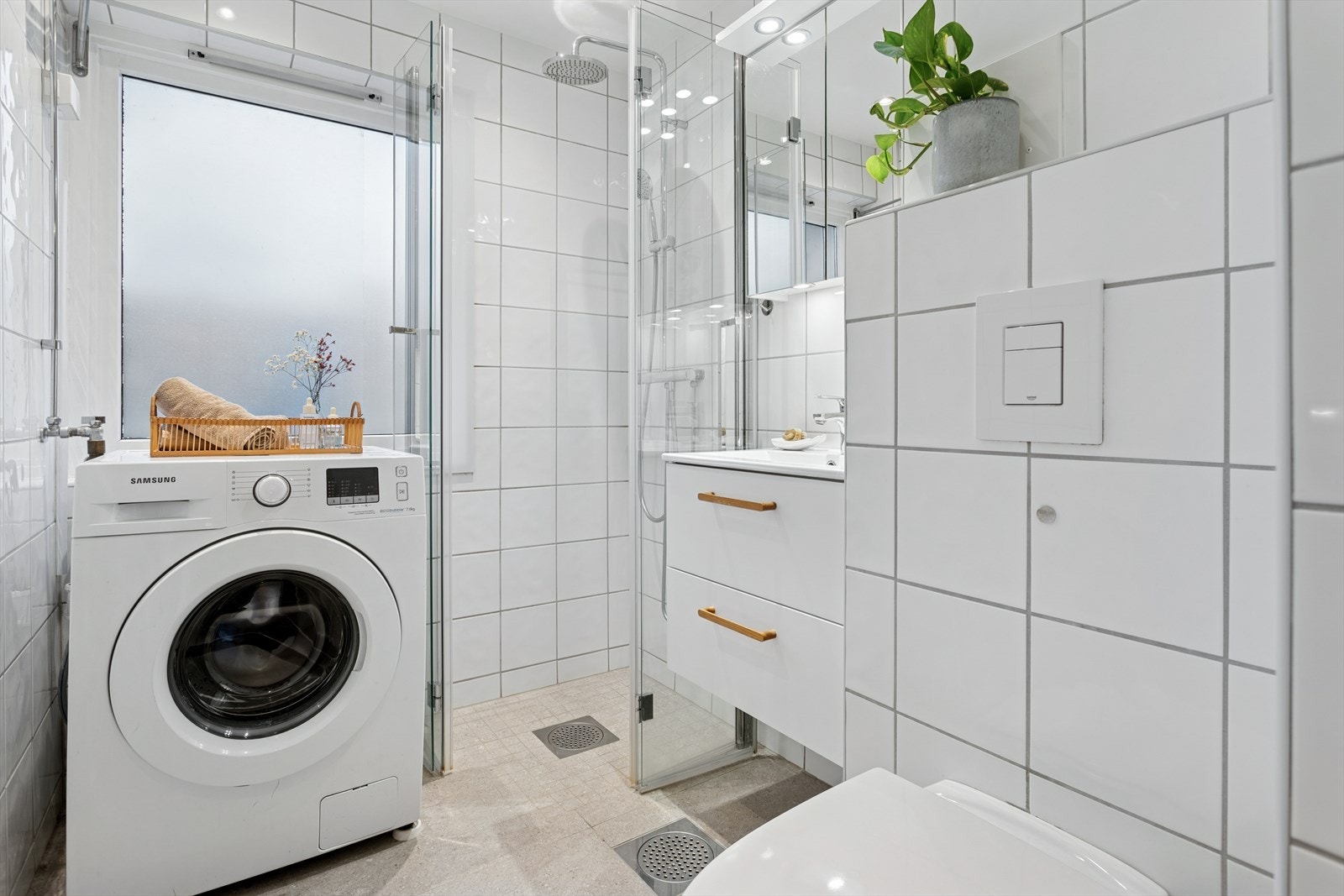 Badet fremstår som delikat og funksjonelt, med fliser på gulv og vegger som gir et lettstelt uttrykk. Rommet er innredet med servantskap i hvit, glatt utførelse med heldekkende servant, samt overskap med speildører som gir godt med oppbevaringsplass. Galleribilde