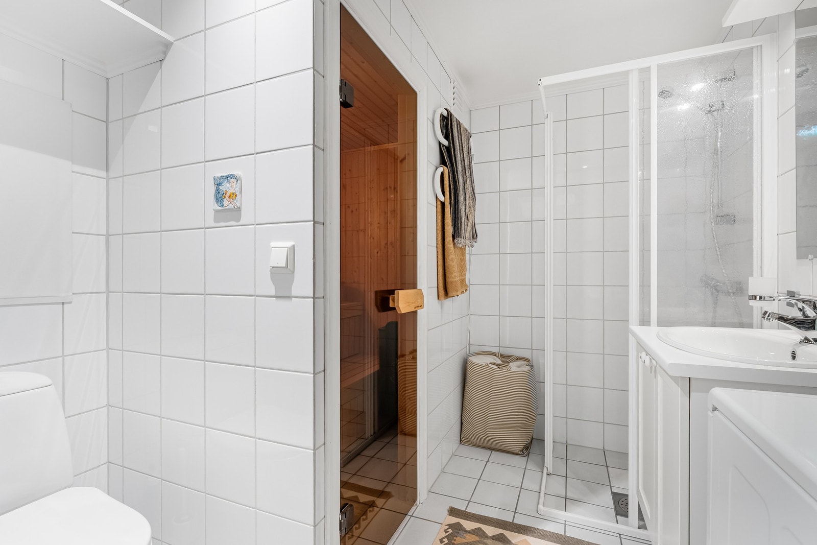 Badet har lyse fliser på gulv og vegger, elektrisk gulvvarme, downlights og mekanisk ventilasjon. Videre er rommet innredet med baderomsinnredning med servant, stort innfliset speil, toalett og dusjnisje Galleribilde