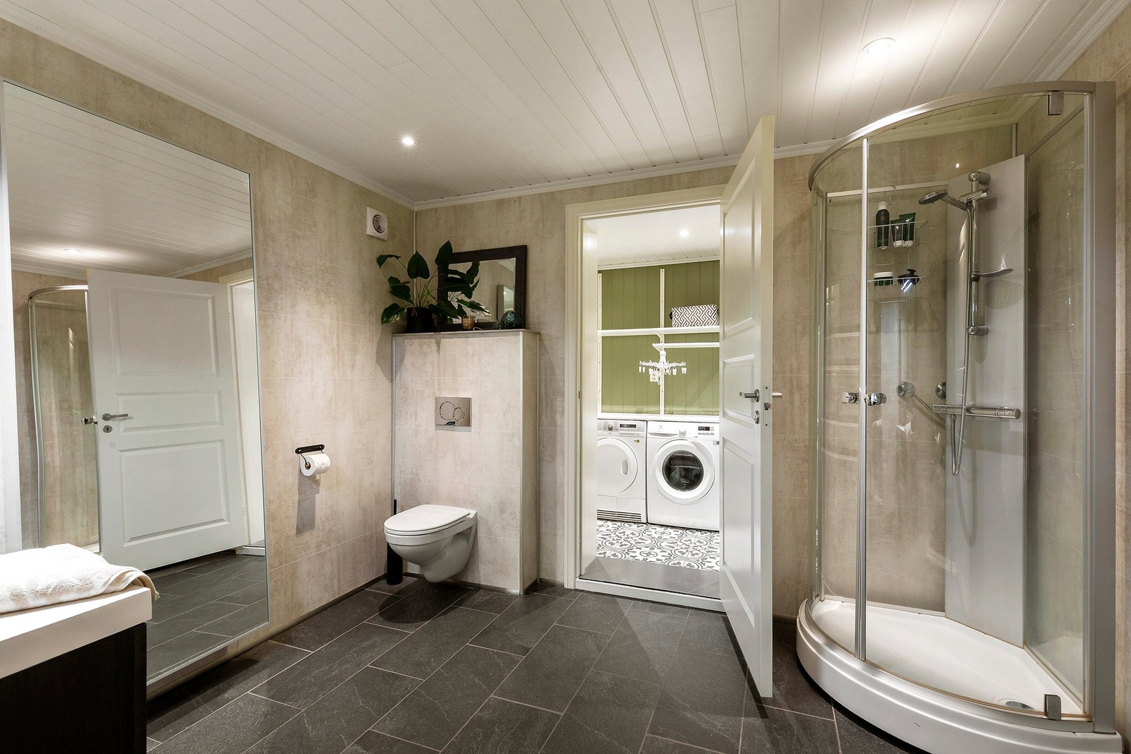 Innbydende baderom med påfølgende vaskerom i sokkel. Det er flis på gulv, gulvvarme, våtromsplater på vegger, panel og downlights i himling. Galleribilde