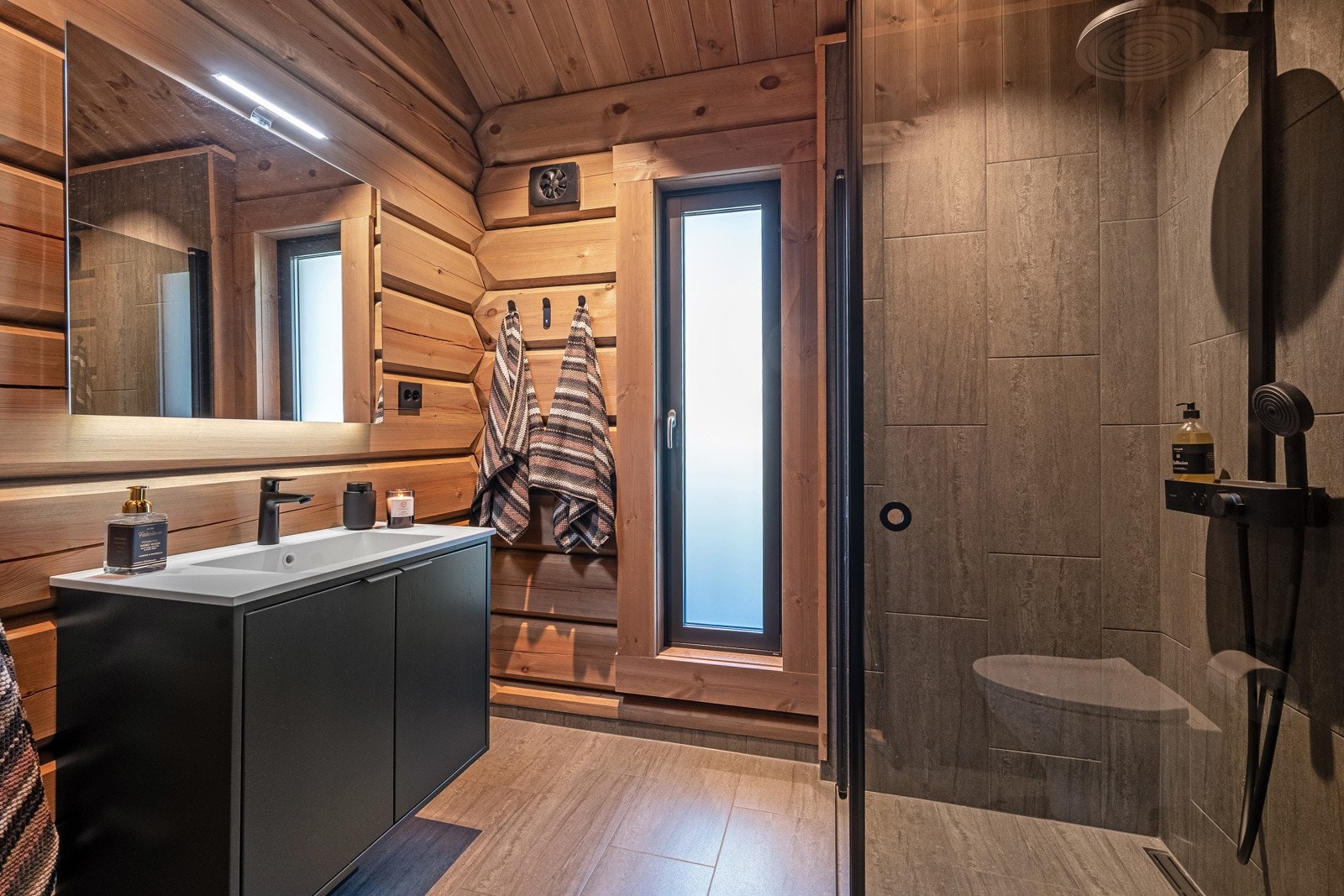 Badet ved entreen er også meget lekkert og har nesten den samme utførelsen med delikate fliser på gulvet og veggene i dusjsonen, kombinert med tømmervegger og en vegg kledd med stående panel fra Vyrk. Galleribilde