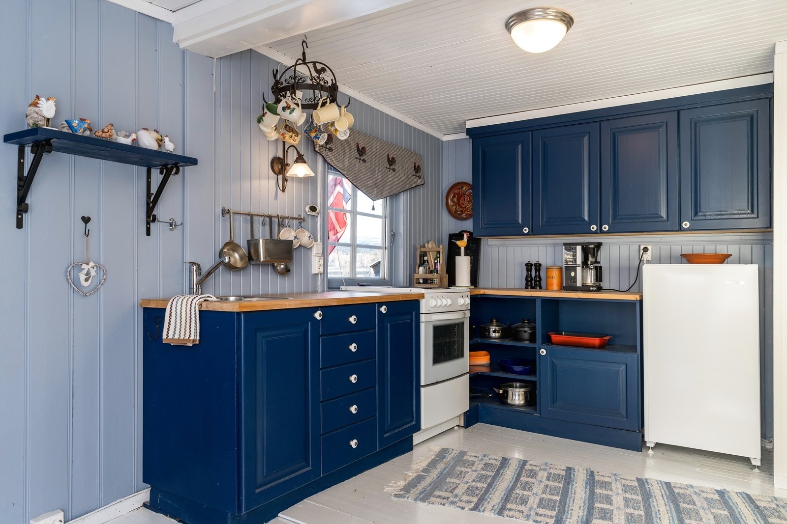 Kjøkken og innredning malt i sjarmerende blåtoner. Kjøkkenet er innredet med komfyr, kjøleskap og vask. Hytta fikk innlagt vann og avløp i 2025. Galleribilde
