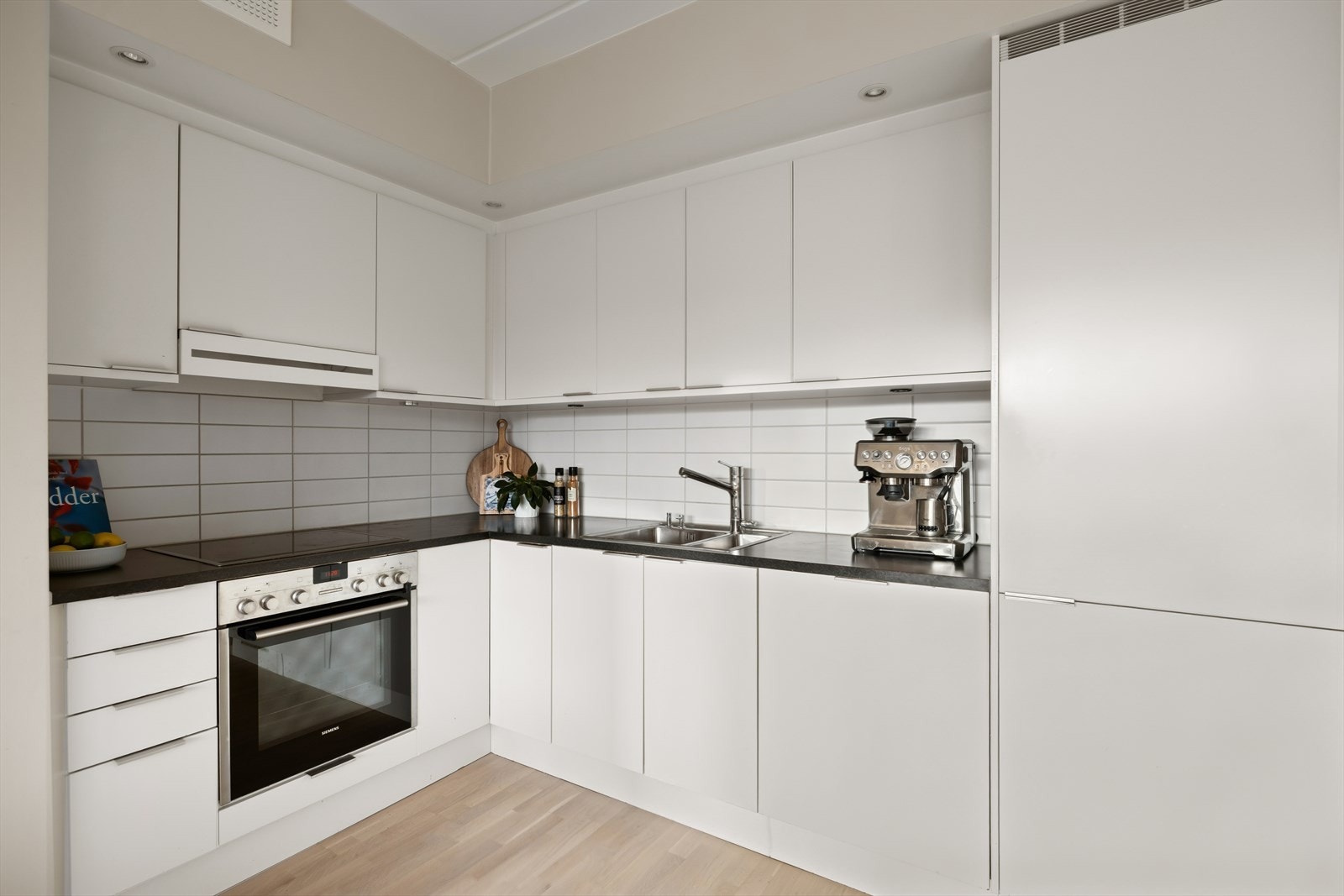 Lyst kjøkken med hvit, glatt innredning og en solid laminat benkeplate. Alt du trenger er integrert: induksjonstopp, stekeovn, oppvaskmaskin, samt kjøl- og fryseskap. Det er satt opp lys under overskapene og fliser mellom skapene for en fin finish. Galleribilde