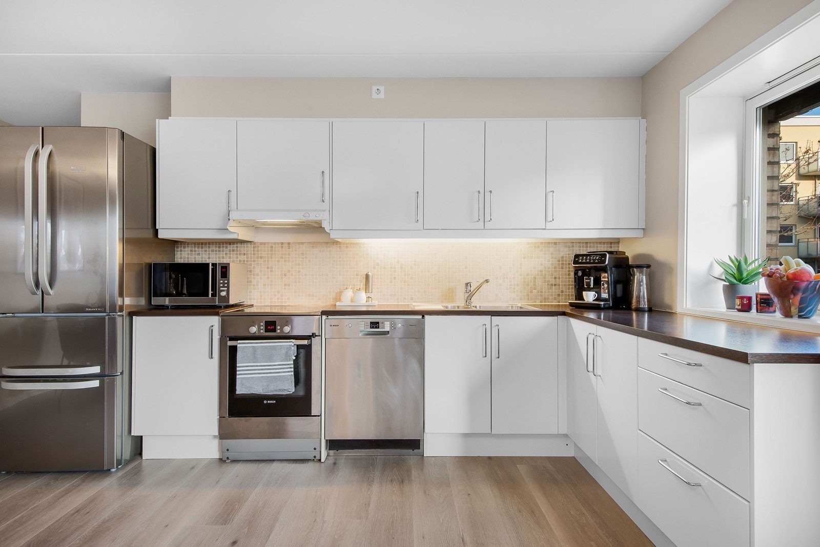 Kjøkkenet er funksjonelt plassert i et hjørne, med mosaikkflis over benk, tidløs innredning og plass til hvitevarer. Galleribilde