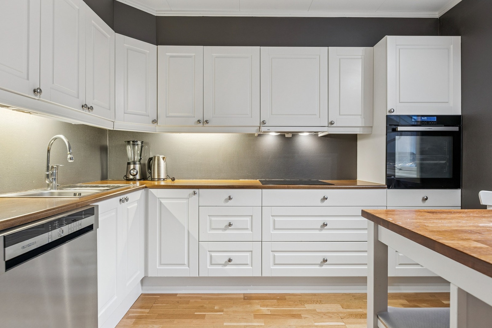 Kjøkkeninnredningen har hvite profilerte fronter som gir et tidløst uttrykk, kombinert med laminert benkeplate som er både slitesterk og lett å holde ren, samt integrert komfyr. Galleribilde