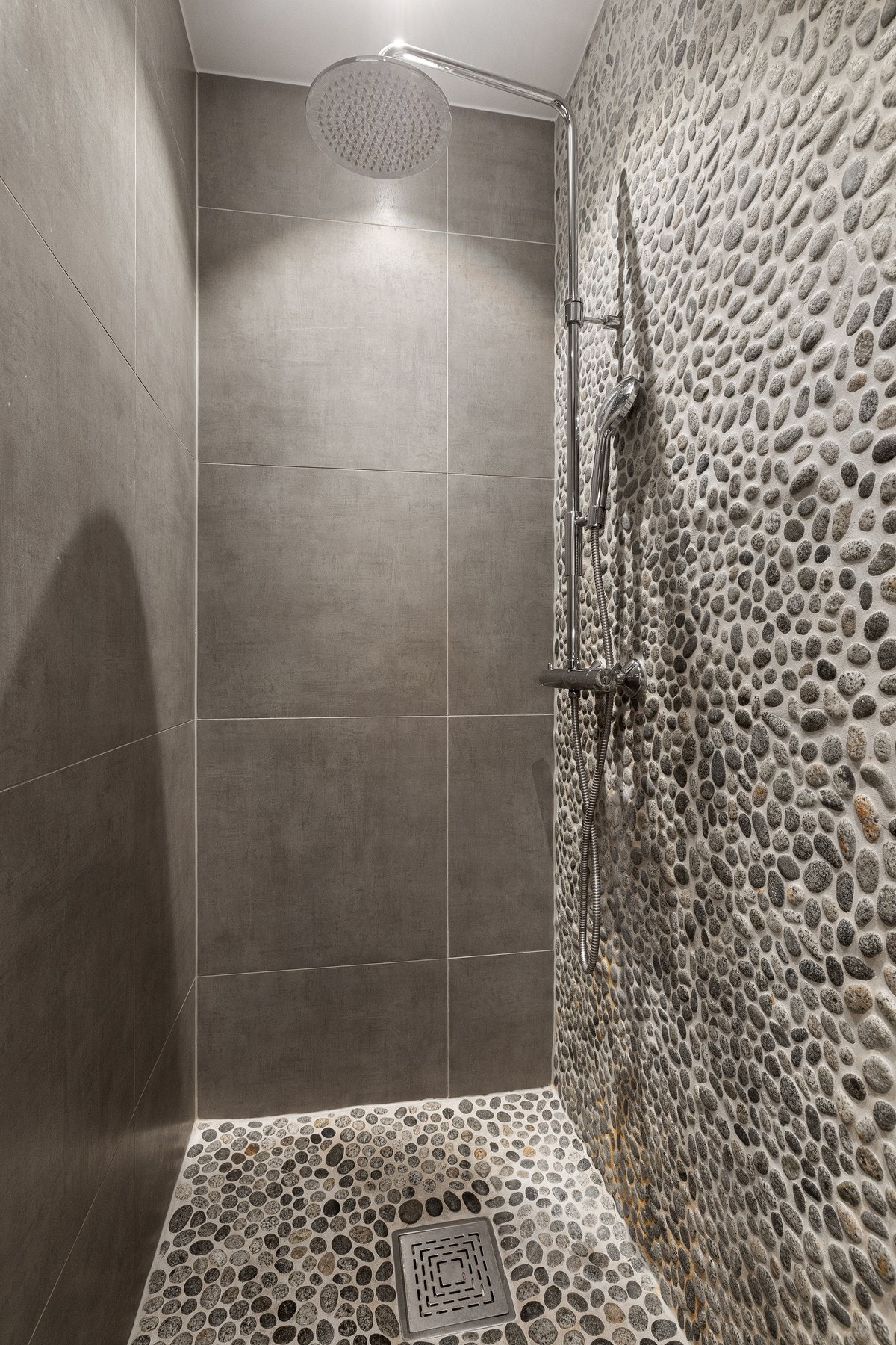 Vegg- og gulvflater er belagt med store, grå fliser som gir et rolig og tidløst preg, mens dusjsonen er fremhevet med elvestein som tilfører rommet karakter og dybde. Galleribilde