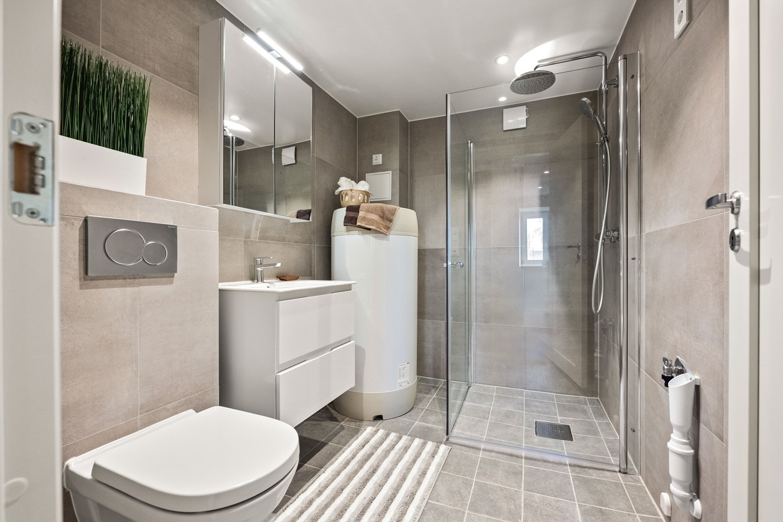 Pent flislagt bad med varmekabler i gulv. Hvit baderomsinnredning med servant, speilskap, toalett og dusjhjørne med regndusjarmatur. Opplegg for vaskemaskin. Galleribilde