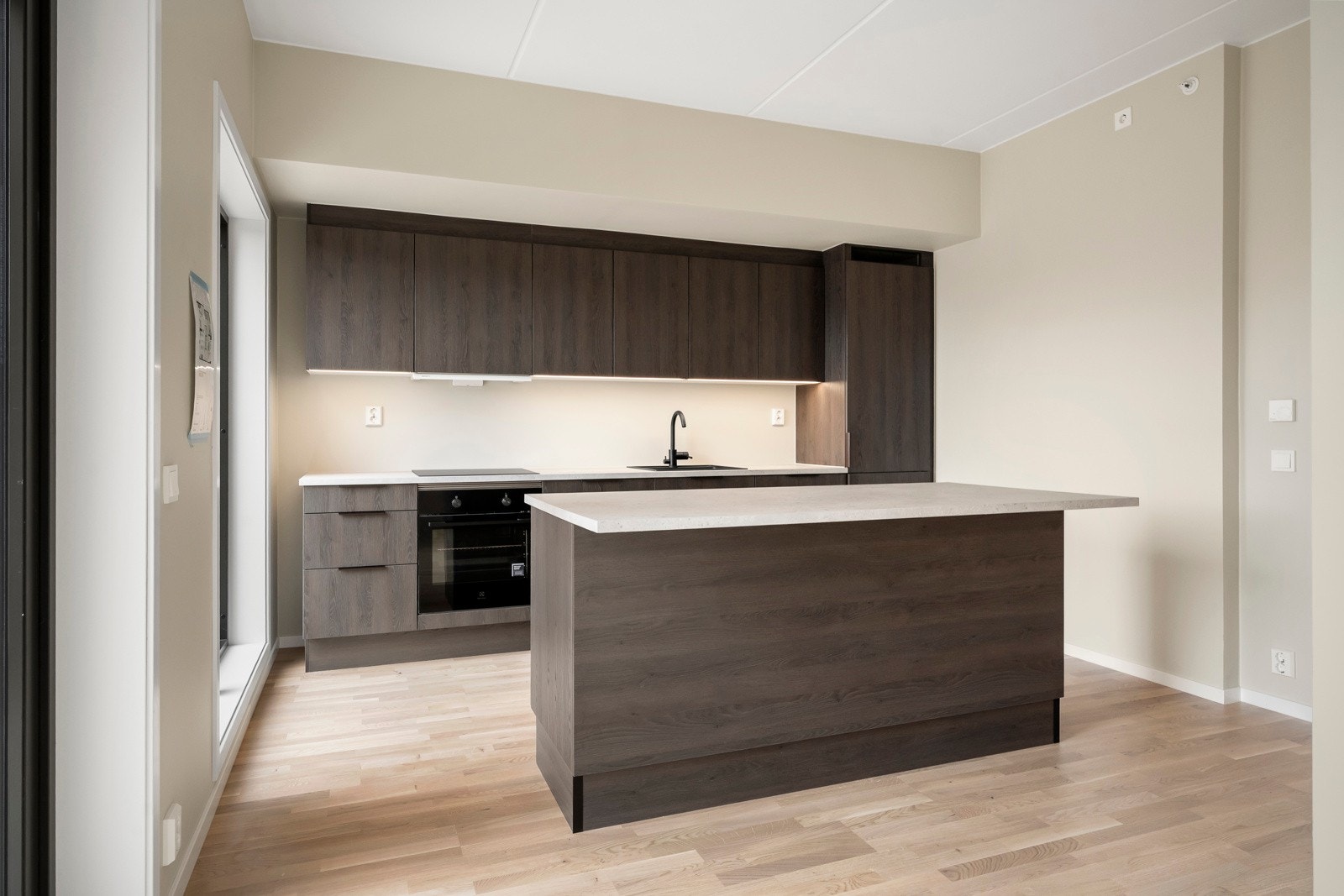 Stilrent kjøkken fra HHT med stor kjøkkenøy, oppgradert og påkostet utover opprinnelig standard. Kjøkkenet har Mood-fronter, håndtak i eloxert brun, sort nedfelt vask, blandebatteri Evo 184 i matt sort, og laminert benkeplate i fargen Sandsten. Galleribilde