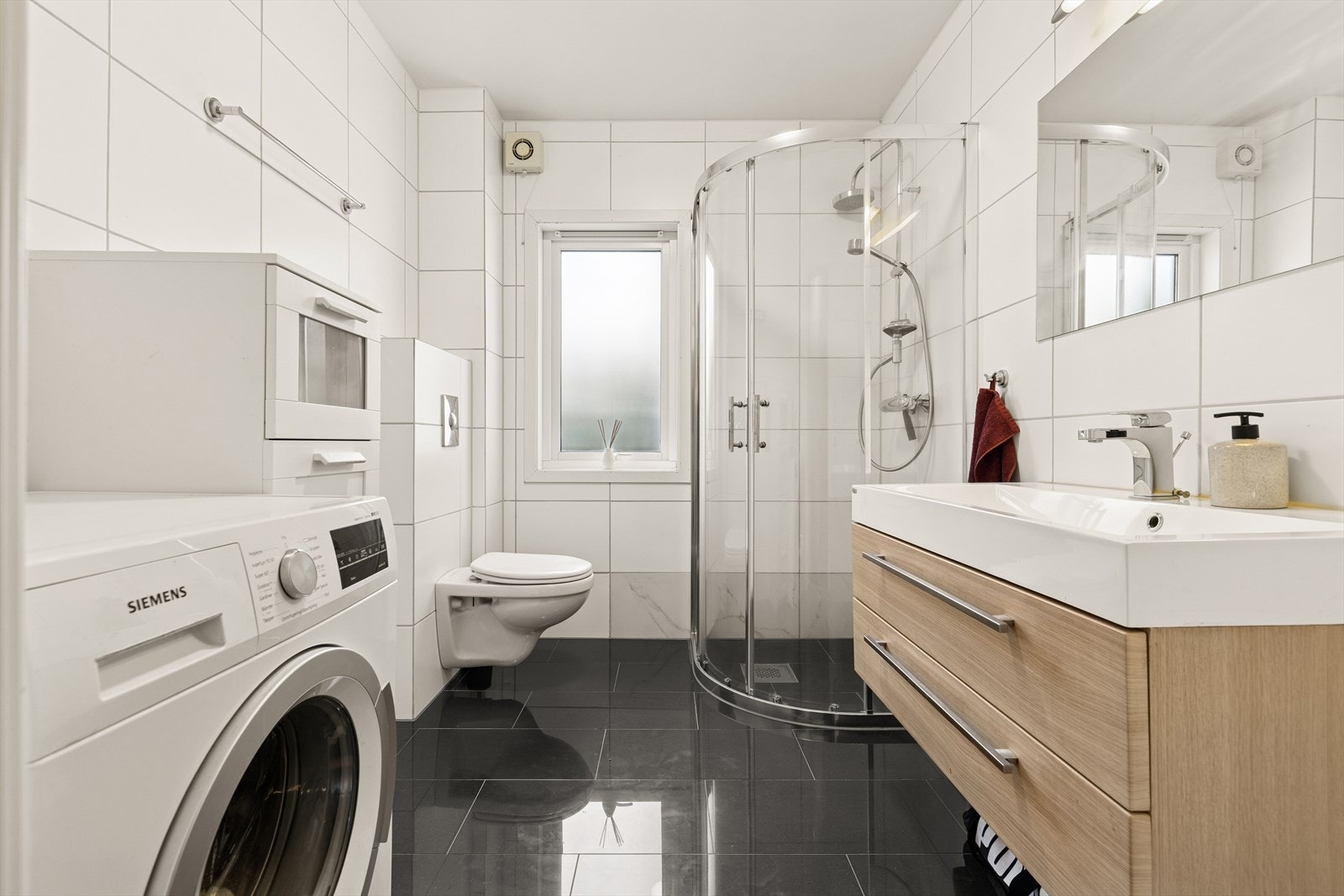 Badet er flislagt og innredet med lys baderomsinnredning, dusjsone, vegghengt toalett samt opplegg for vaskemaskin og tørketrommel i høyden. Et tidløst og funksjonelt bad. Galleribilde