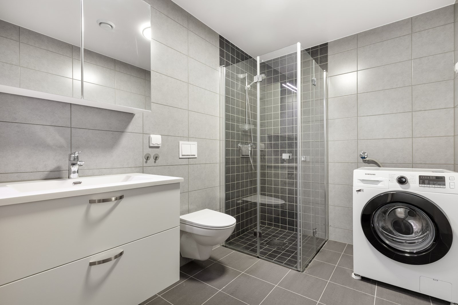 Flislagt bad med mørke gulvfliser, grå veggfliser og opplegg for vaskemaskin. Galleribilde