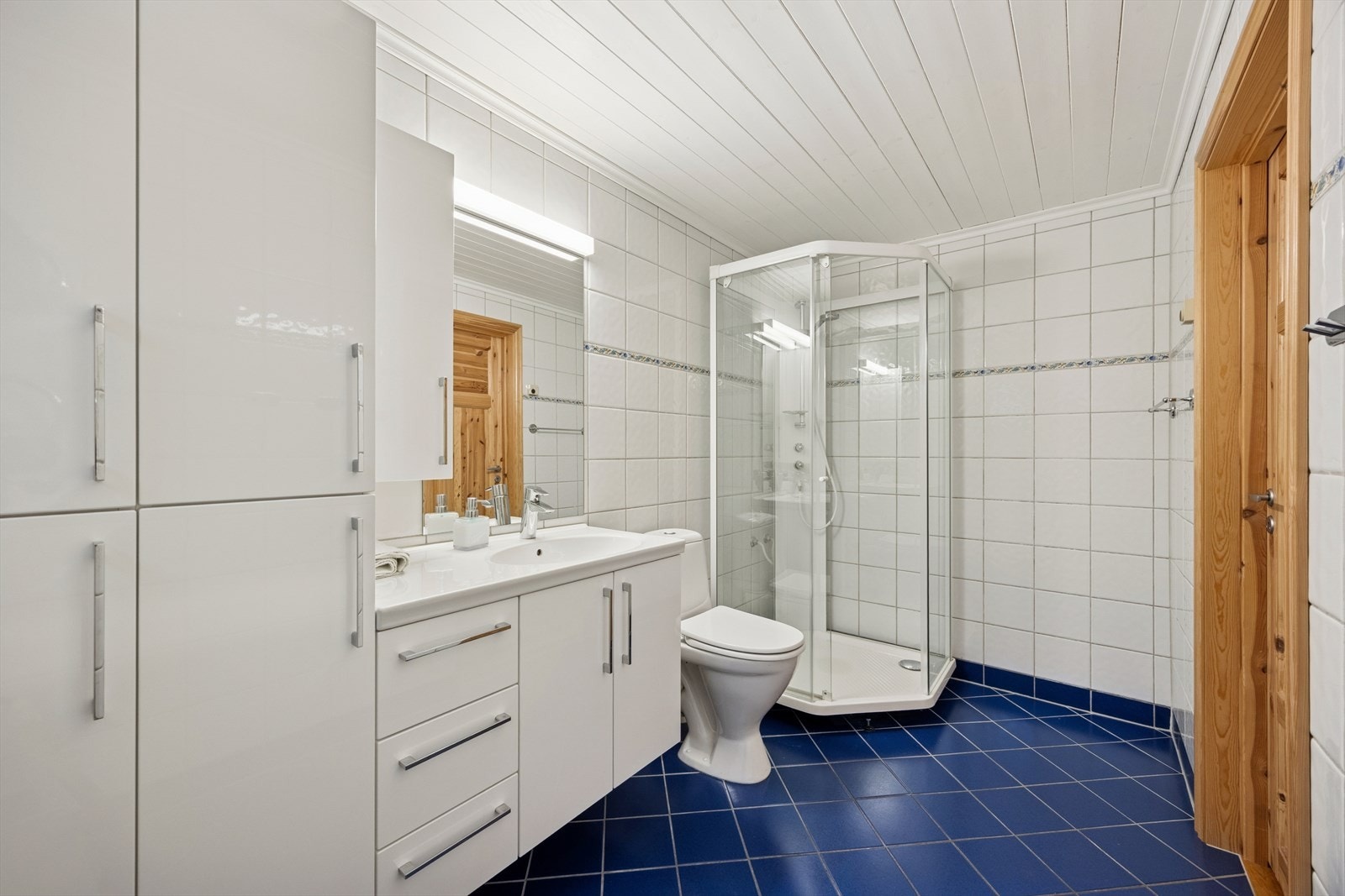 Badet er utstyrt med gulvmontert wc, dusjkabinett, baderomsinnredning med heldekkende servant og speil på vegg. Galleribilde