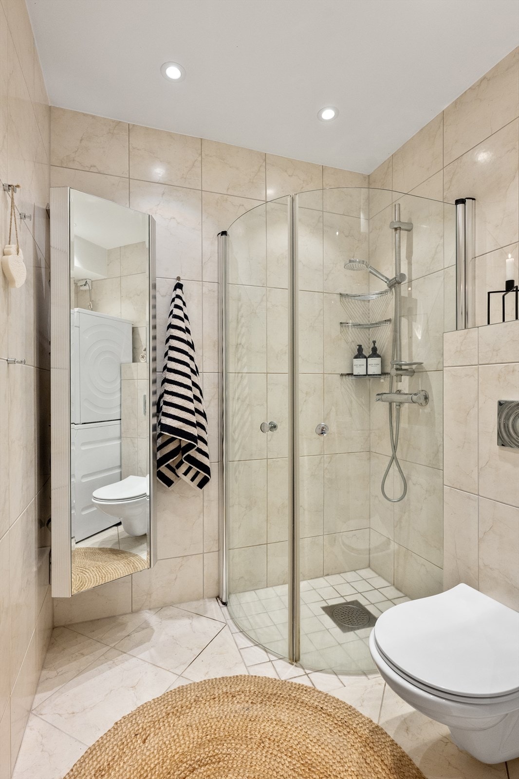 Badet er innredet med dusj på gulv, veggfestet toalett, servant med benkeskap, overskap og veggskap med speilfronter. Galleribilde