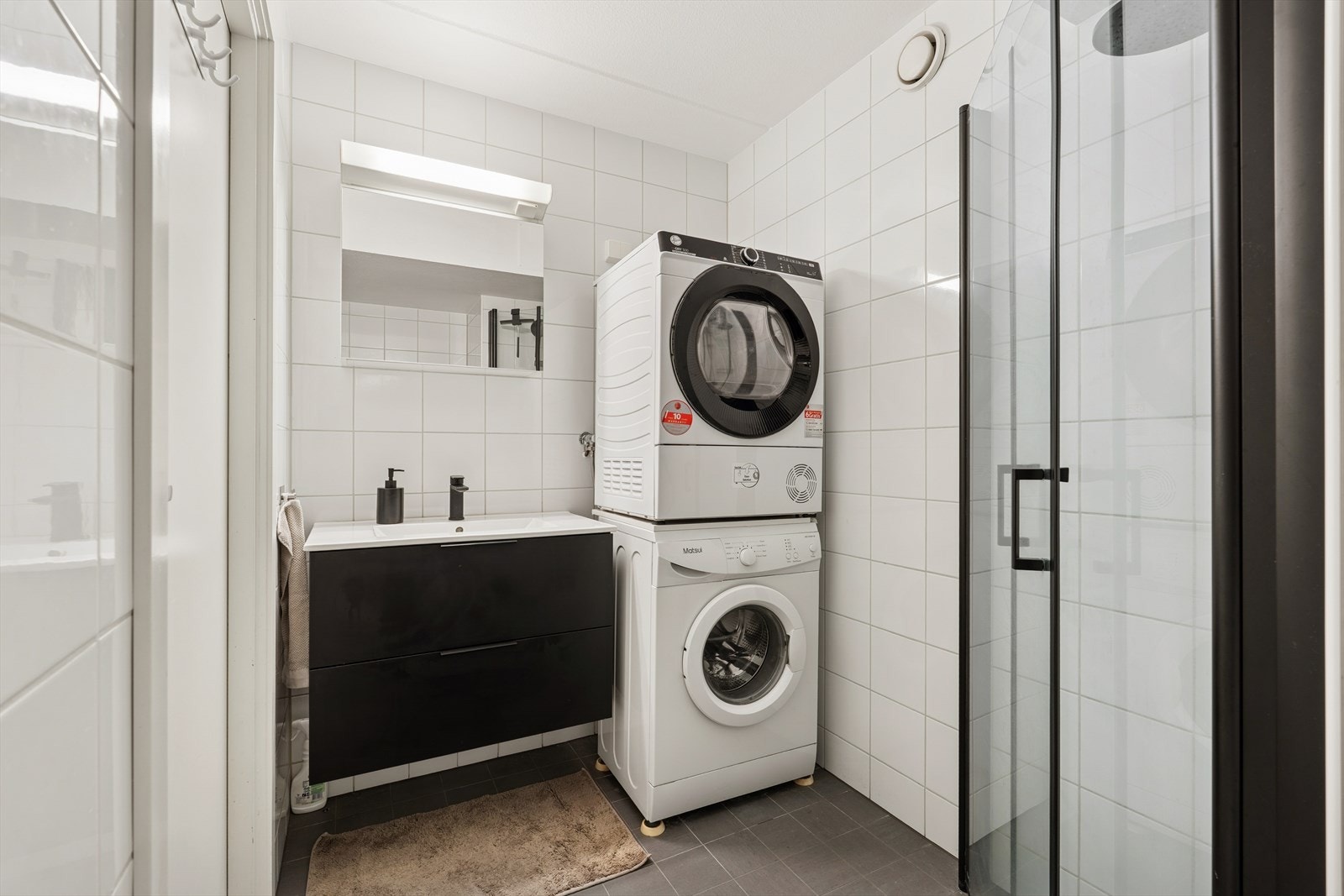 Det er fliser på gulv og vegger, dusjhjørne med glassdører og opplegg for vaskemaskin. Galleribilde