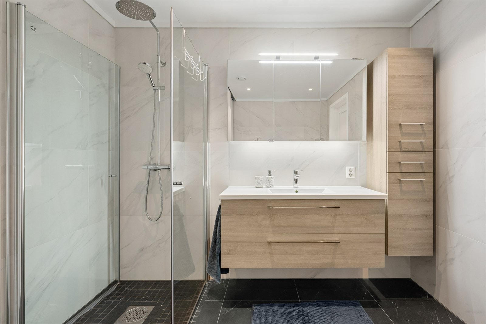 Badet har flislagt gulv med integrerte varmekabler og fliser på veggene. Rommet er utstyrt med en innredning med nedfelt servant, vegghengt speilskap, vegghengt toalett og et dusjhjørne med glassvegger. Galleribilde