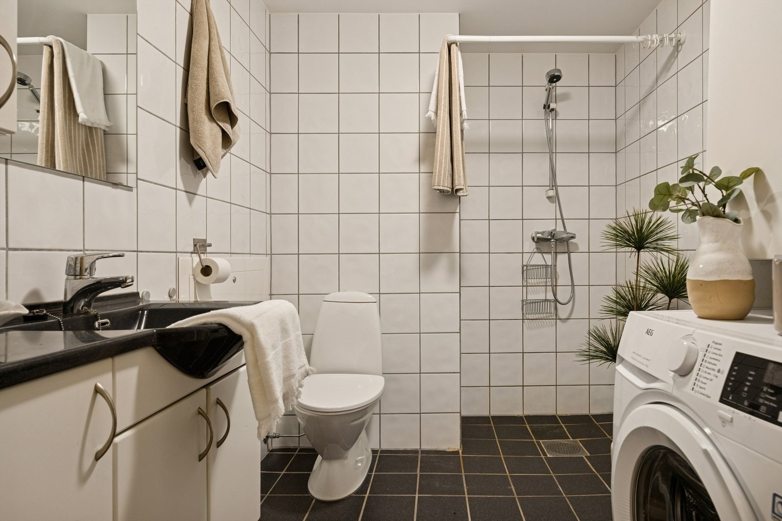 Flislagt baderom med en servantinnredning, gulvmontert toalett og en dusjnisje. Opplegg for vaskemaskin. Vaskemaskin medfølger i handelen. Galleribilde