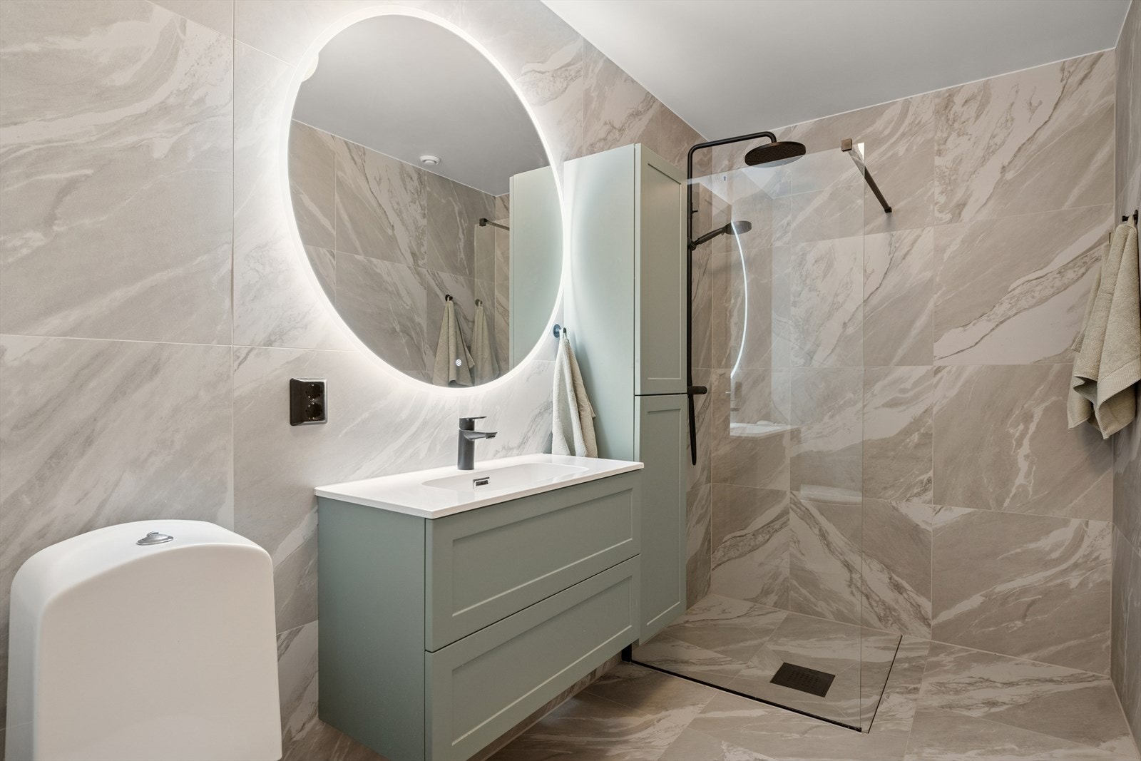 Innredningen består videre av en heldekkende servantplate på lys skuffeseksjon, veggskap i samme stil, rundt speil med belysning og toalett. Rommet har lyse og mønstrede fliser på gulv og vegger i en eksklusiv utførelse. Galleribilde