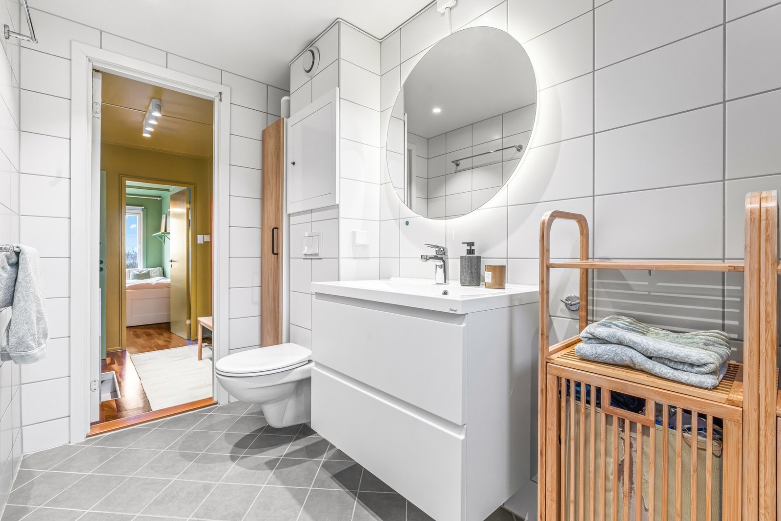 Baderomsinnredningen består av heldekkende servant med hvite skuffer, et hendig høyskap og et rundt speil med bakenforliggende belysning. Galleribilde