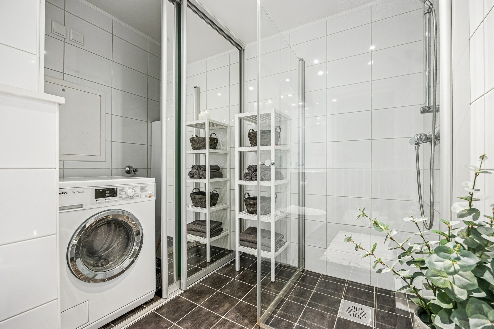 Badet er flislagt med gulvvarme og har servant, speil med overlys, dusjhjørne med dører og vegghengt toalett, samt en praktisk løsning med integrert skap for vaskemaskin. Galleribilde