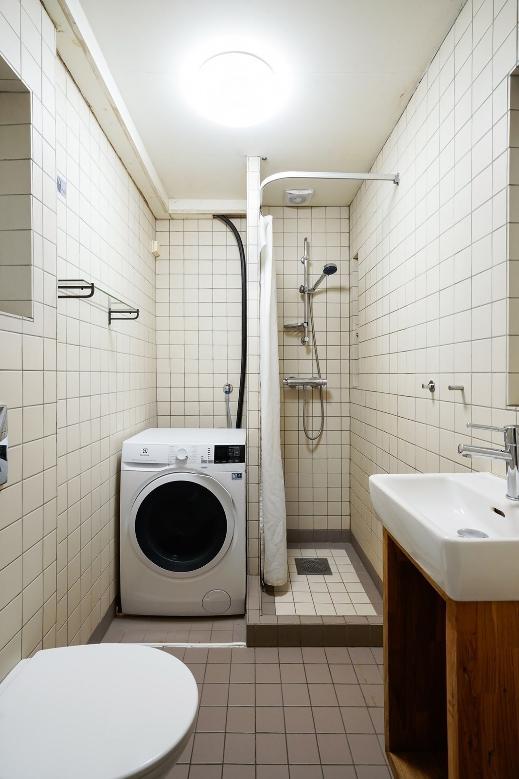 Eldre bad med flis på gulv og flis på vegger. Badet er utstyrt med dusjhjørne med forheng, veggmontert toalett, hvit servant med blandebatteri fra Damixa. Galleribilde