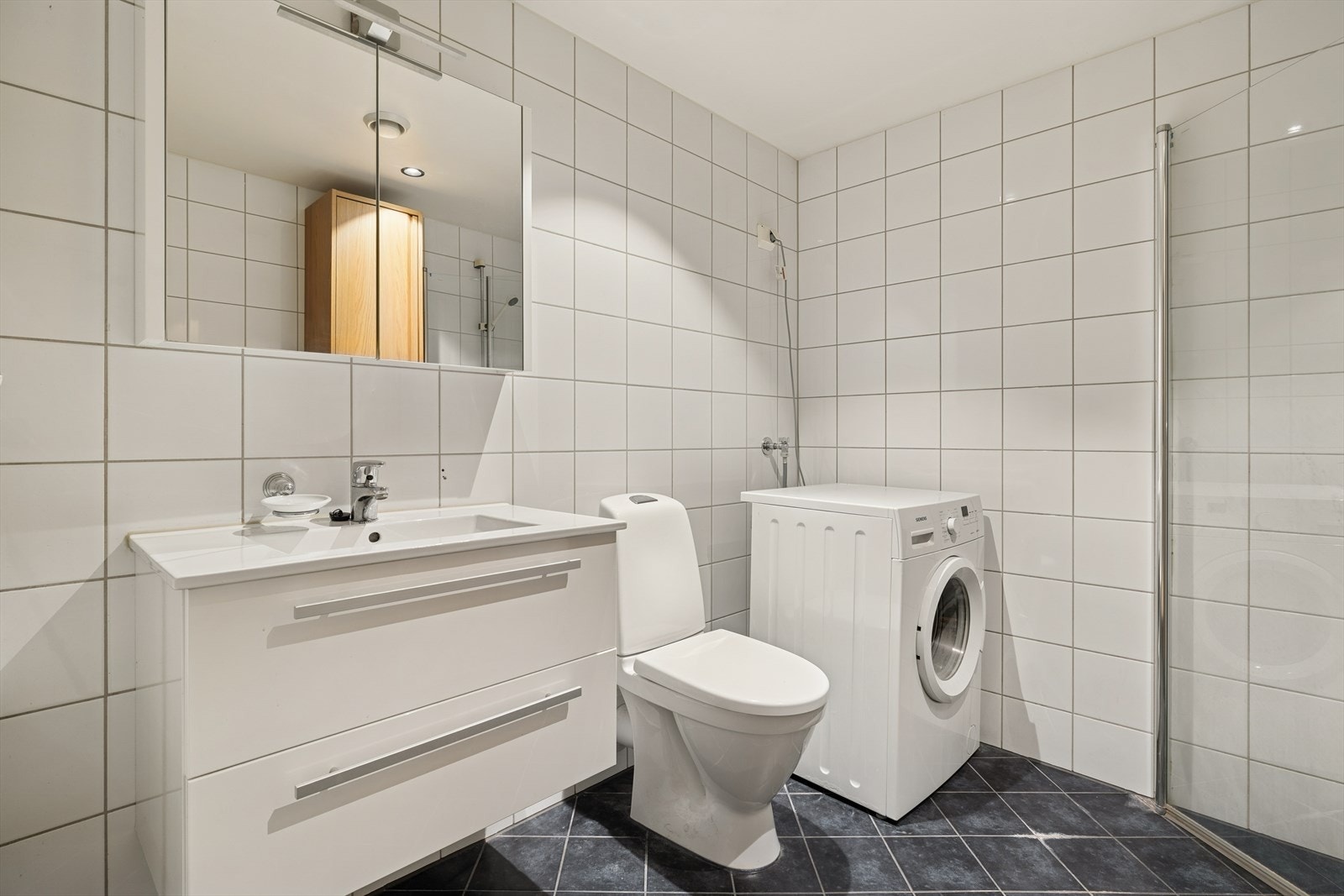 Badet har innredning med nedfelt servant, gulvfestet toalett, dusjhjørne og opplegg for vaskemaskin. Galleribilde