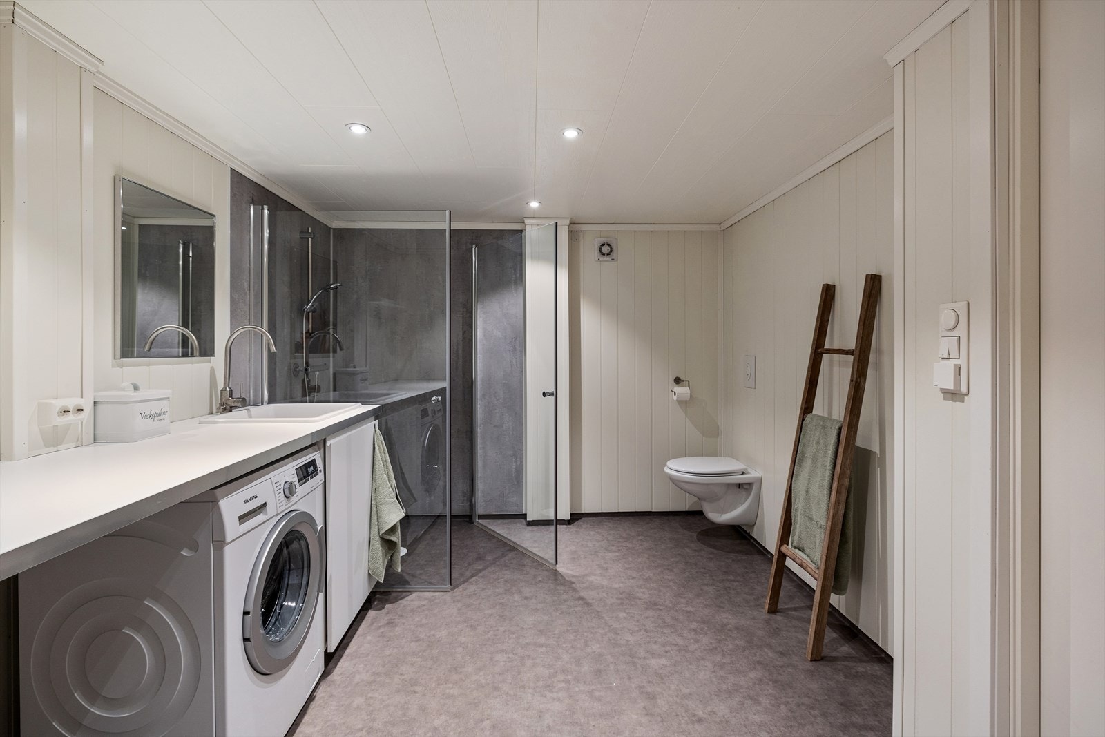 Badet har Baderomsplater og panelplater på veggene og belegg på gulv med gulvvarme. Badet har servant med underskap, speil med tilhørende belysning, dusjvegger, vegghengt wc og opplegg for vaskemaskin. Galleribilde