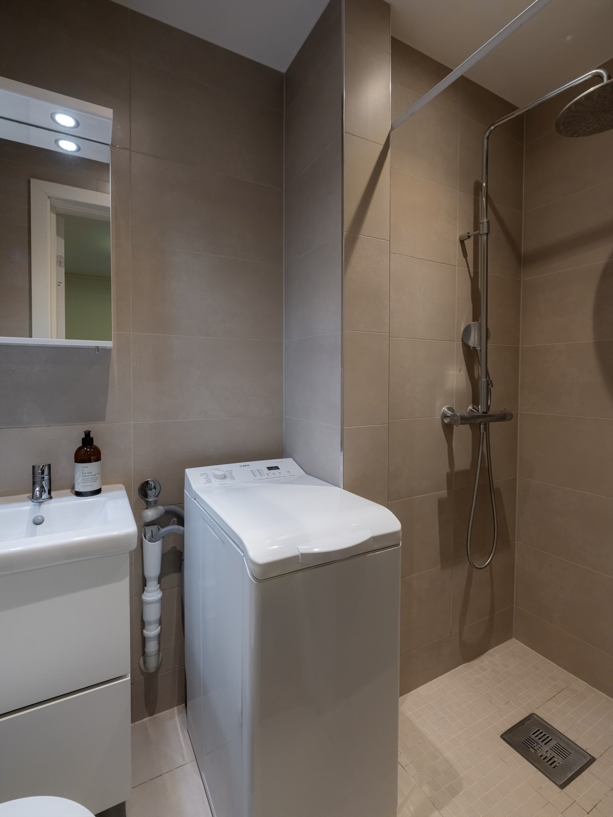 Badet i 1. etasje er flislagt fra byggeår og har moderne innredning med benkeskap med glatte fronter, veggskap med speilfronter, vegghengt toalett, dusjnisje med regnfallsdusj og opplegg for vaskemaskin. Galleribilde