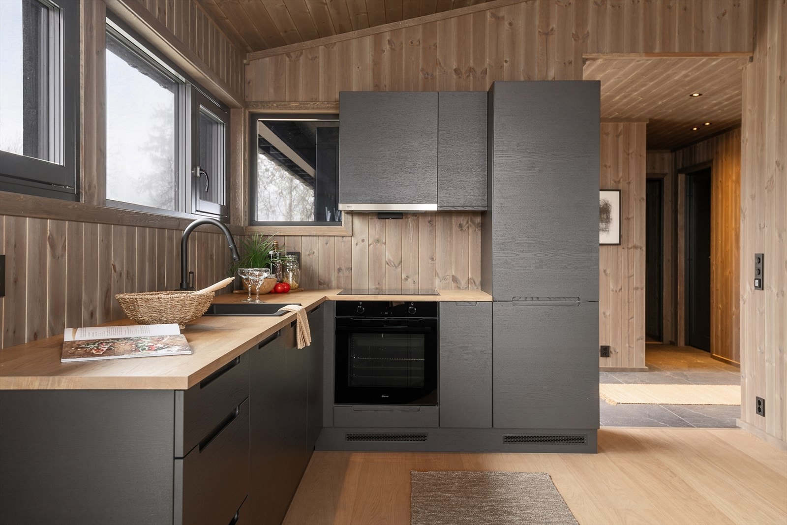 Moderne kjøkken med stilrene, mørke fronter og naturlige tregulv. Lysinnslipp fra store vinduer gir en innbydende atmosfære.. Illustrasjonsbilde fra Svartskardvegen 66. Denne hytta er speilvendt i forhold til avbildet hytte. Galleribilde