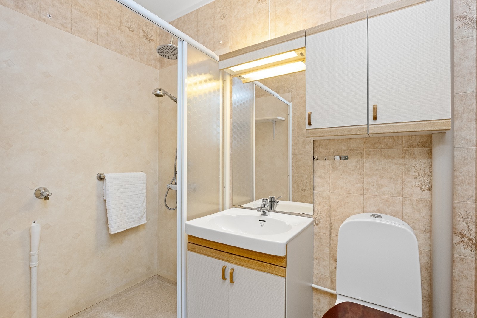 Eldre bad med belegg på gulvet og våtromstapet på vegger, samt servantinnredning med overhengende speil og skap. Dusjen er plassert i egen nisje, og badet ventileres via ventil i himling. Galleribilde