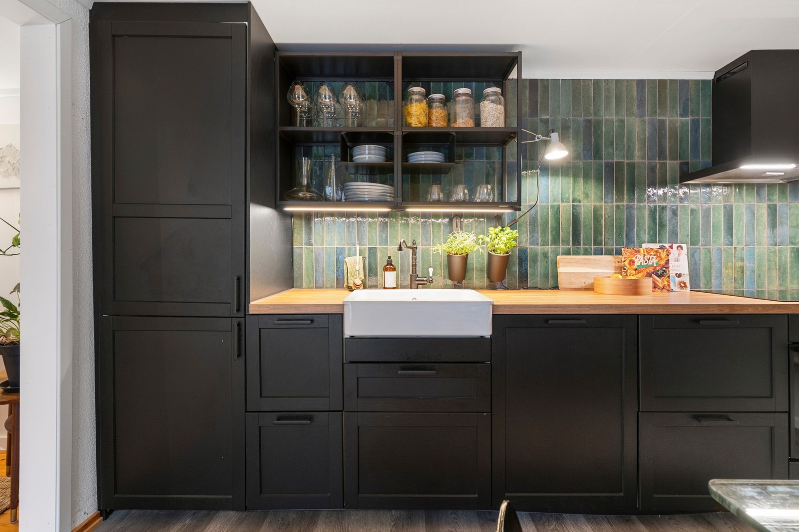 Kjøkken med mørke skapfronter, trebenk og grønne fliser. Hyller over vasken gir ekstra oppbevaringsplass. Galleribilde