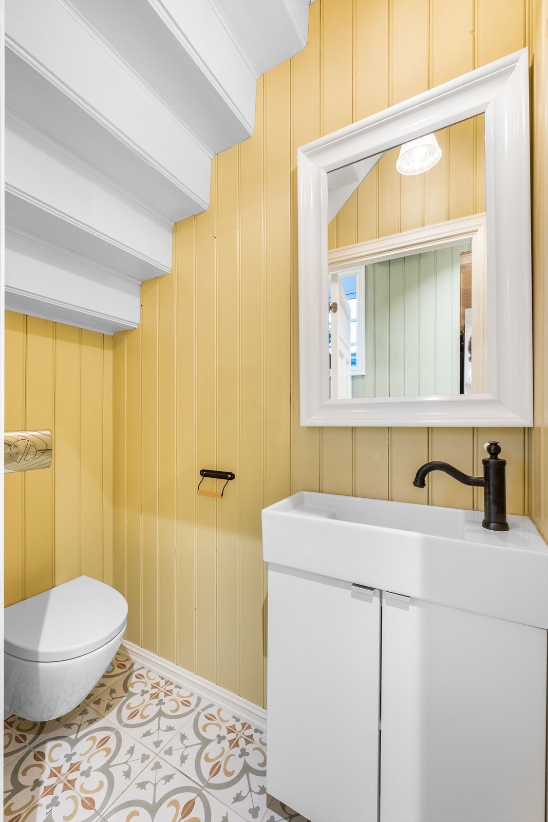 Praktisk toalettrom i 1.etg med flislagt gulv med varmekabler og malt panel i himling og vegger. Rommet er innredet med veggmontert toalett og servant med fabrikklakkerte fronter. Det er speil på vegg over servanten. Galleribilde