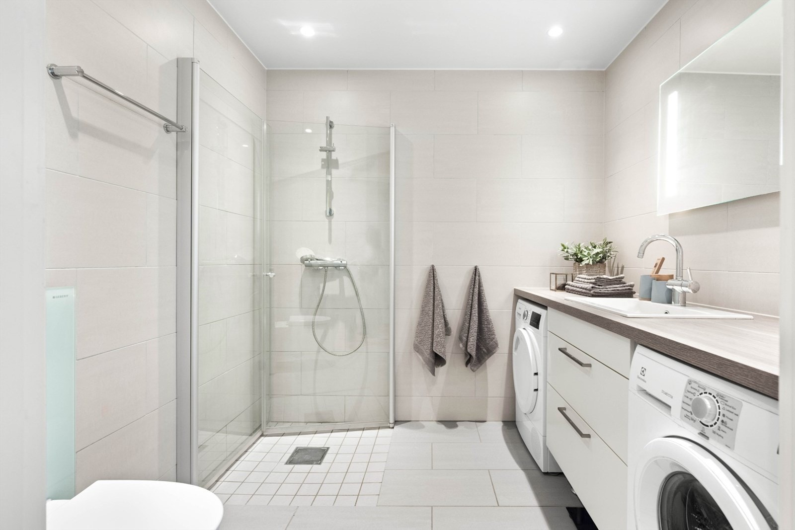 Baderom med lyse fliser på gulv og vegger, med vannbåren varme i gulvet. Nedsenket dusjhjørne med innfellbare dører. Galleribilde