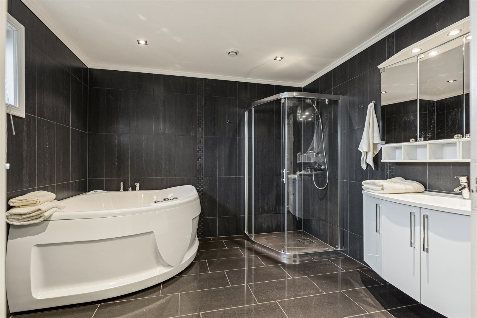 Helfliset baderom innredet med dusjhjørne, servant med skapinnredning og hjørnebadekar. Galleribilde