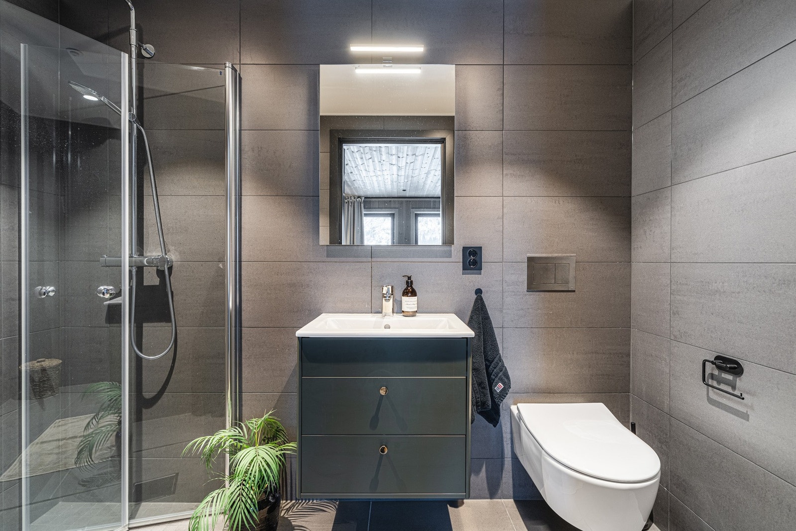 Badet er innredet med servant med underskuffer, toalett, speil med belysning og dusjhjørne med hånddusj og regnfallsdusj. Galleribilde