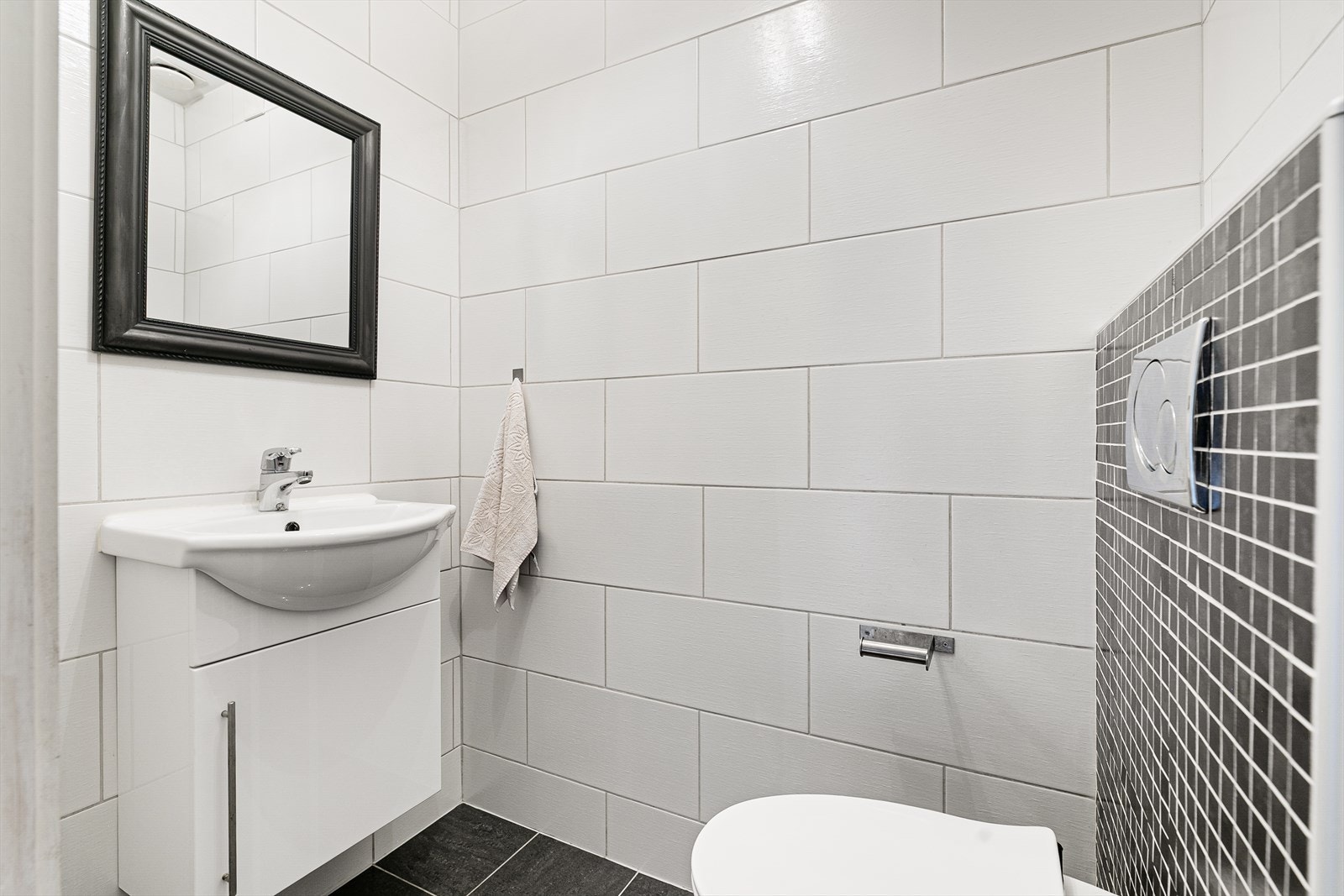 Flislagt toalettrom med vegghengt klosett, servantseksjon med skap og speil. Galleribilde