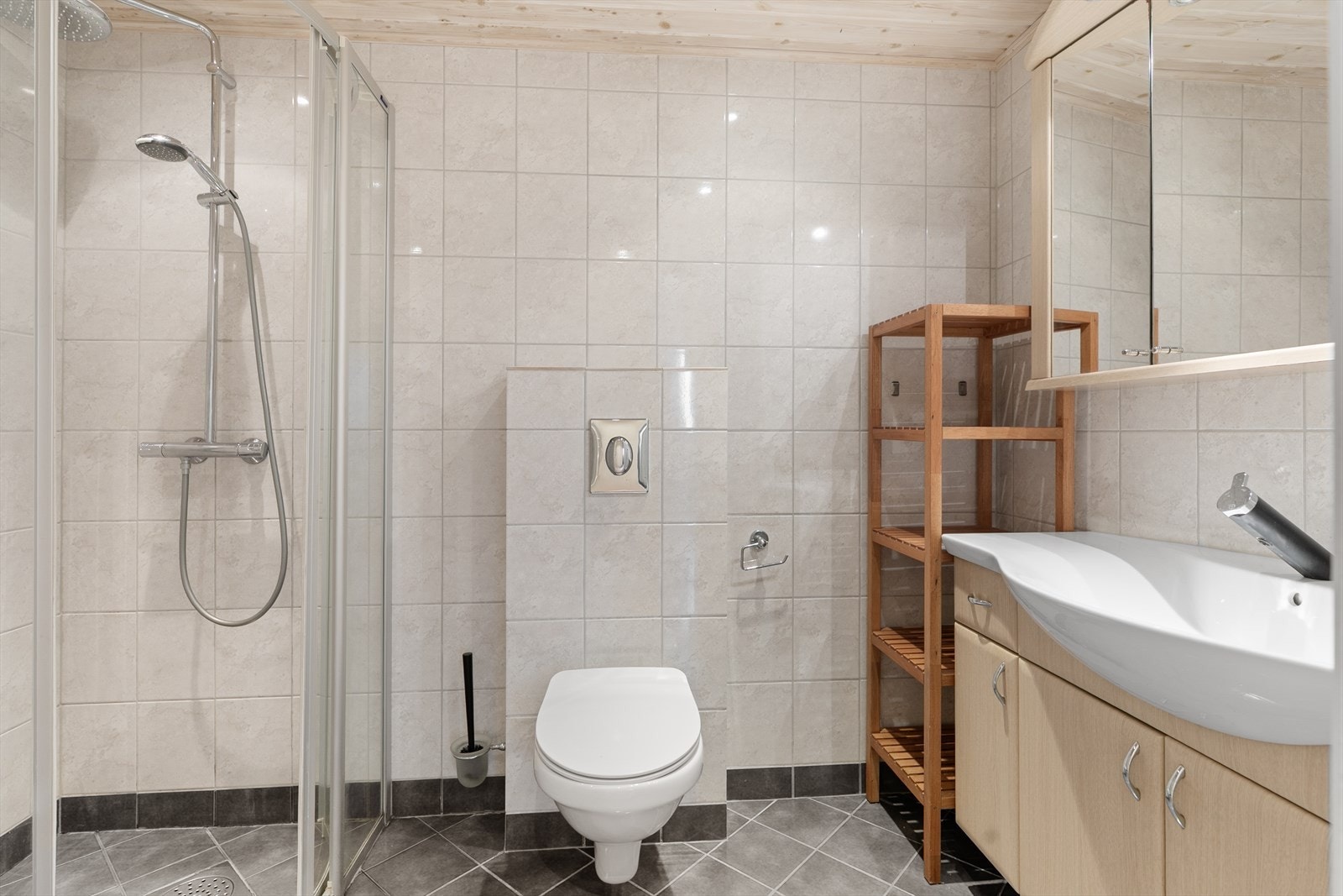 Flislagt baderom inneholdende vegghengt klosett, dusj med inn-/utadslående dører og servantseksjon med skap. Galleribilde