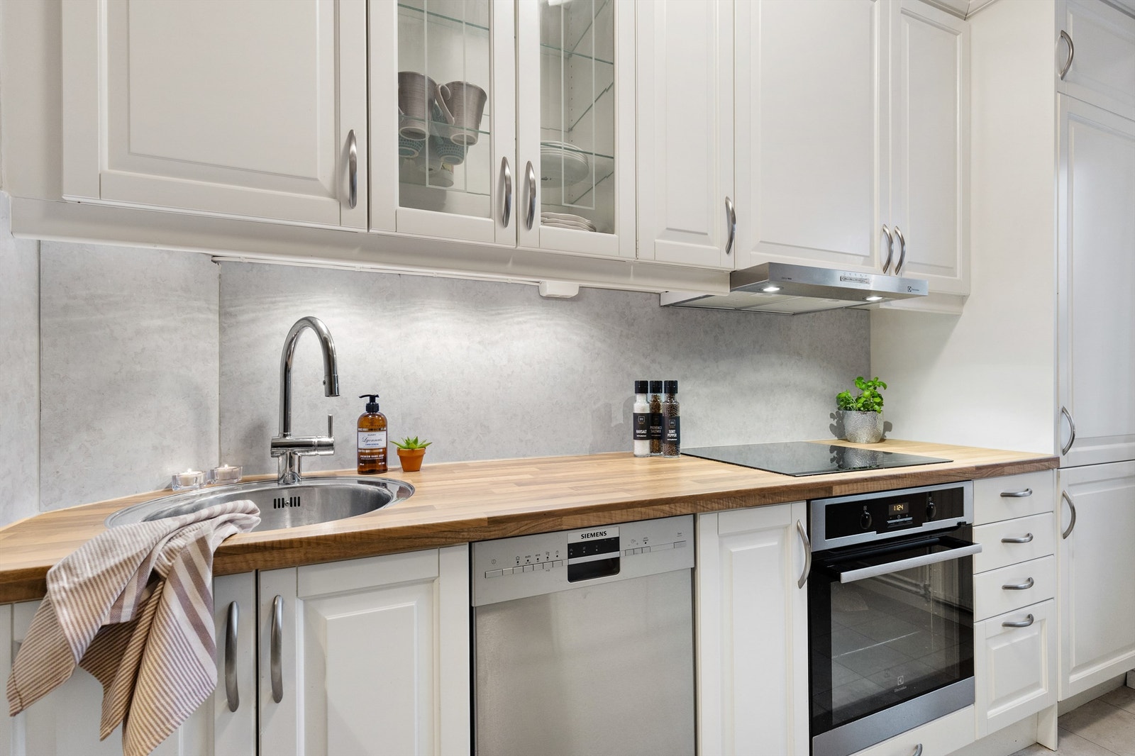 Kjøkkeninnredning med profilerte fronter og laminat benkeplate med nedfelt vaskekum. Integrert induksjonstopp og stekeovn. Det er montert vannstoppsystem og kullfilterventilator over kokeplass. Galleribilde