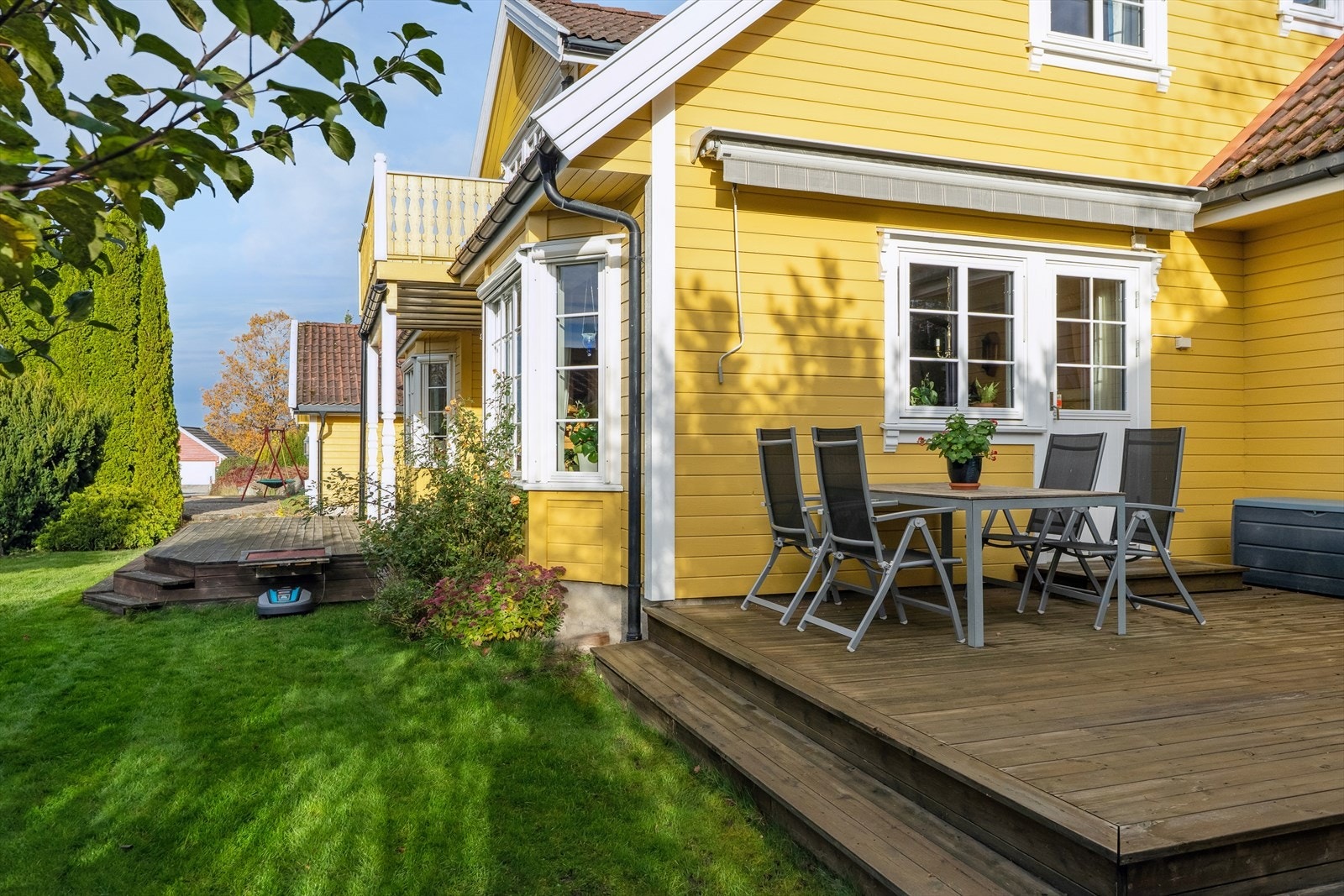 Uteområdet inkluderer en treplatting med plass til utemøbler, omgitt av velstelt hage. Galleribilde