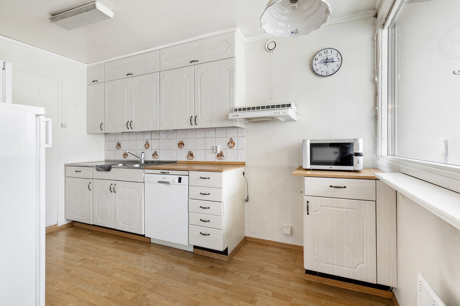 Kjøkkeninnredning med glatte fronter og laminat benkeplate med nedfelt vaskekum og overskap. Det er opplegg for oppvaskmaskin. Fliser på vegg over benkeplaten. Kullfilterventilator over kokeplass. Galleribilde