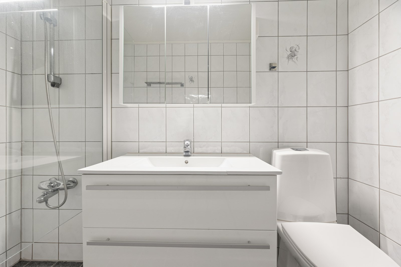 Rommet er innredet med en servant nedfelt i baderomsinnredning, speilskap med overliggende lys, toalett og et dusjhjørne med dusjvegg i glass. Galleribilde