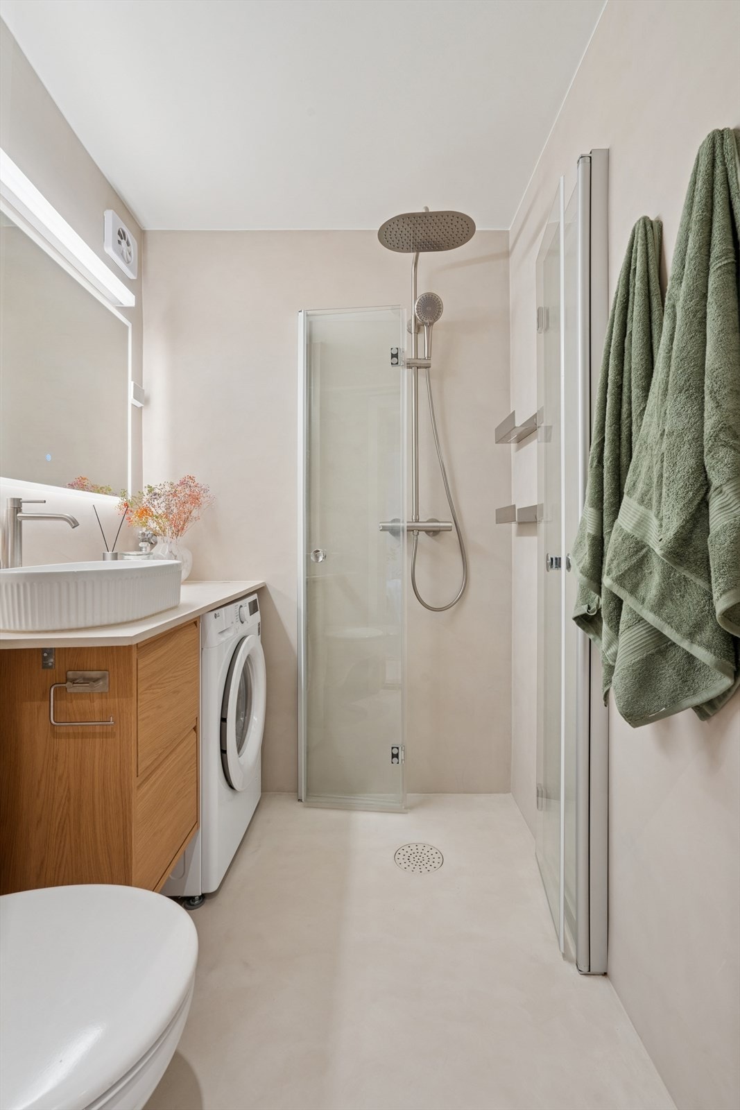 Speil med lys, vegghengt toalett og dusj i hjørnet bak svingbare glassvegger Galleribilde