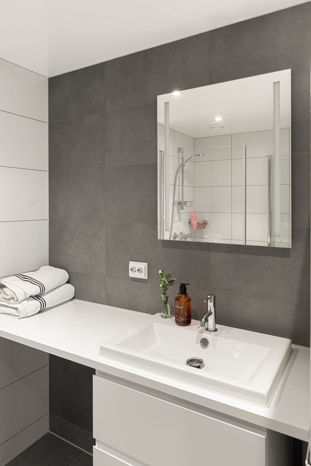 Moderne baderomsinnredning med skuffer og lys laminert benkeplate nedfelt servant, speil med integrert belysning over. Baderommet er også utstyrt med dusjhjørne med innfellbare glassdører og vegghengt toalett. Plass og opplegg for vaskemaskin/tørketrommel Galleribilde