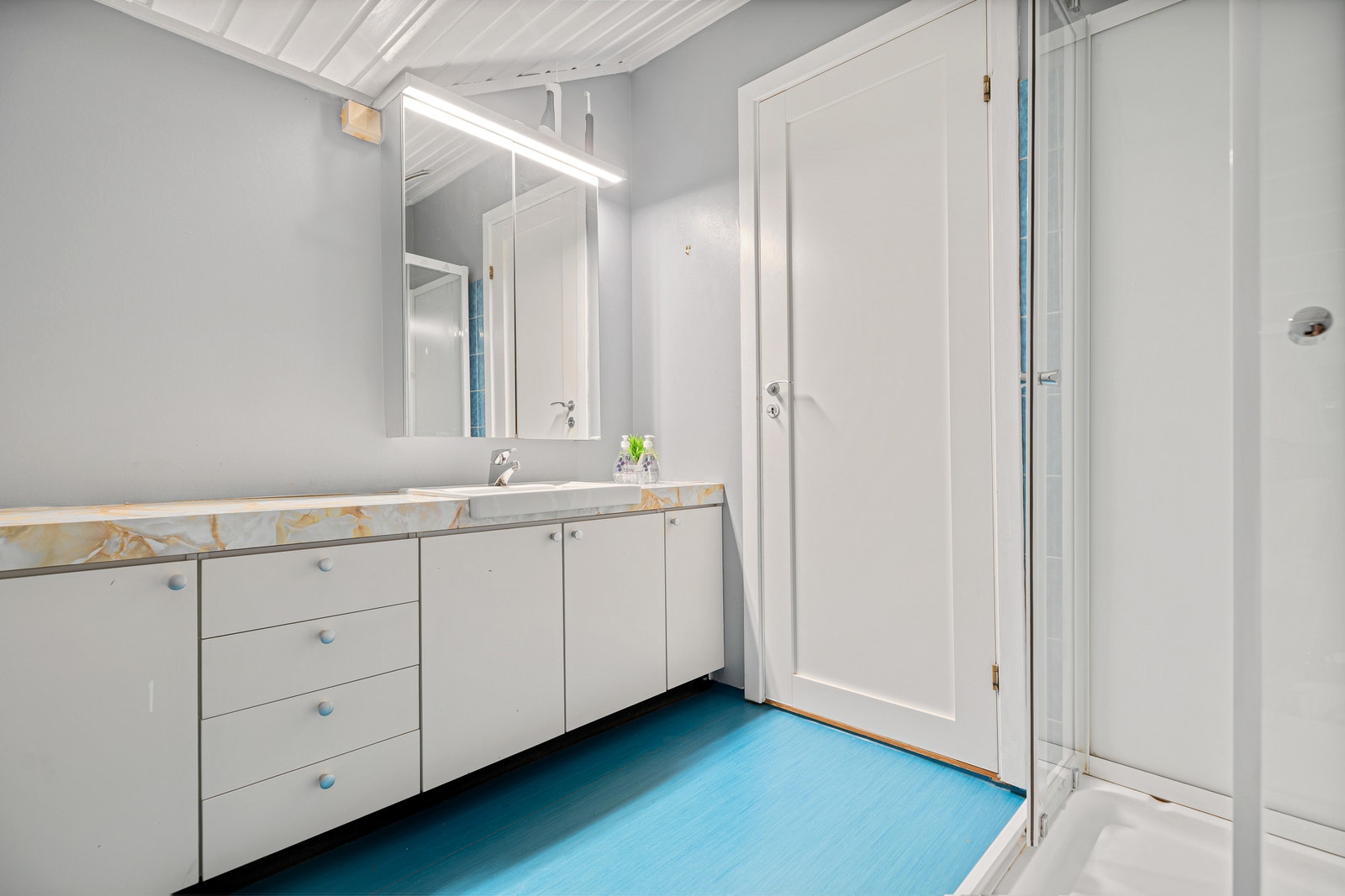 Bad med lang servantbenk, dusjkabinett og blått gulvbelegg. Galleribilde